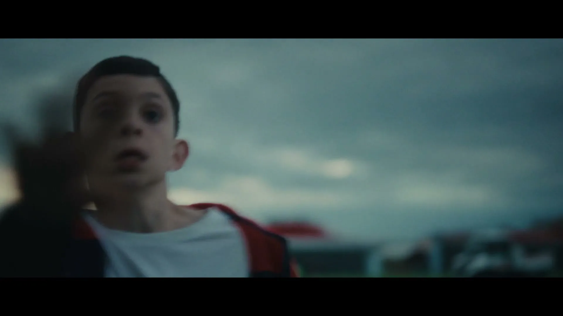 A young boy with dark hair and a red jacket runs across a field under a cloudy sky, looking scared.