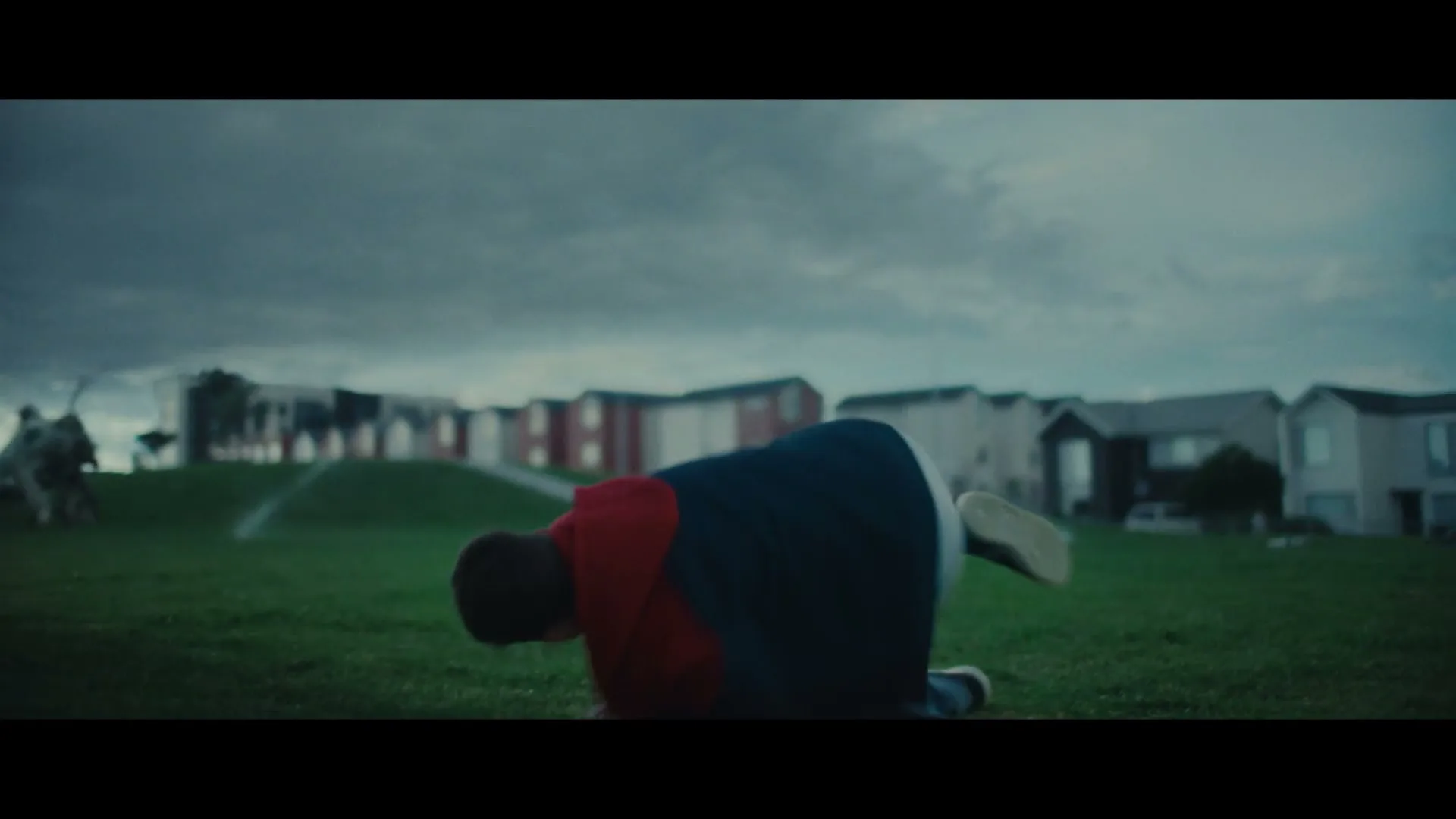 A young boy in a red hoodie and blue pants falls backward onto a grassy field under a cloudy sky, with houses in the background.