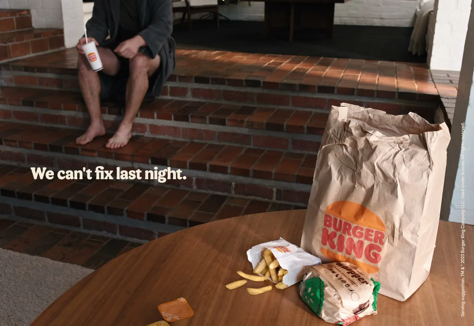 A man in a bathrobe sits on brick steps, looking at a Burger King meal on a wooden table in a home interior.