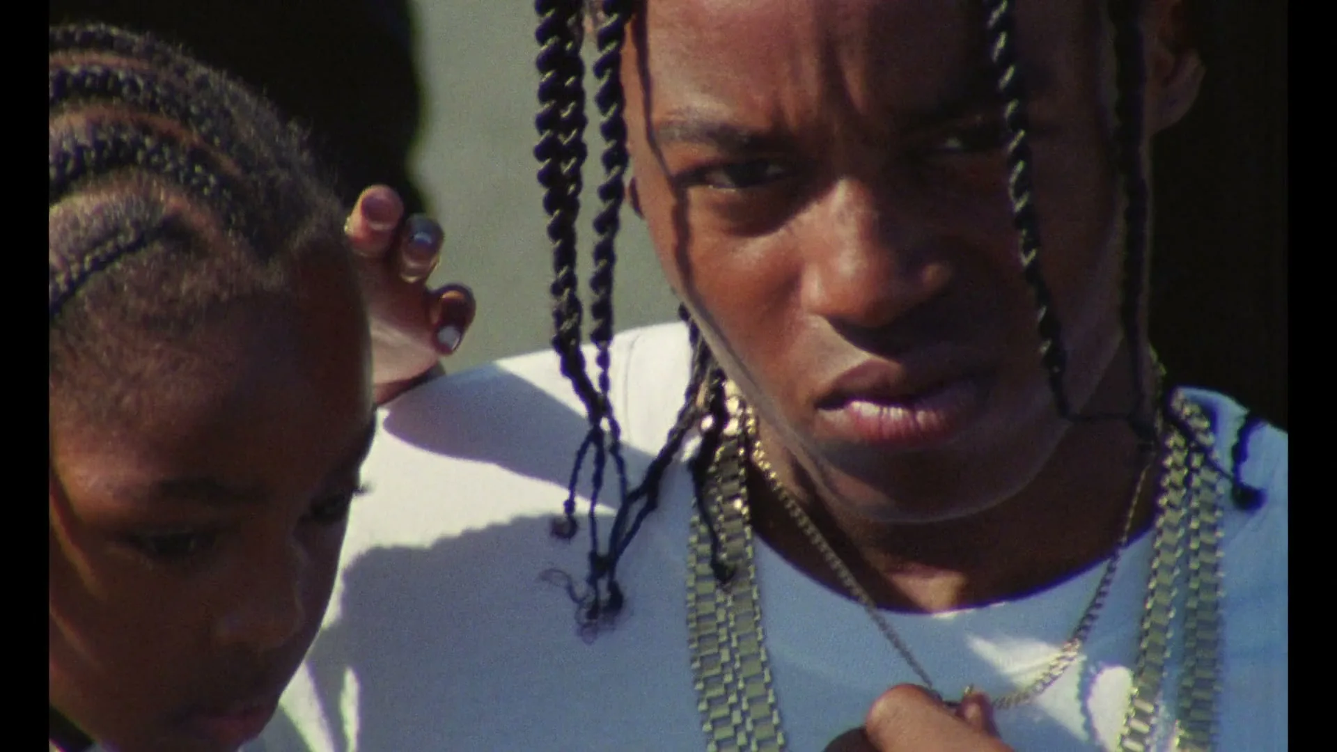 A young Black man with braided hair and gold chains looks down, then up at the camera, with a young Black girl beside him.