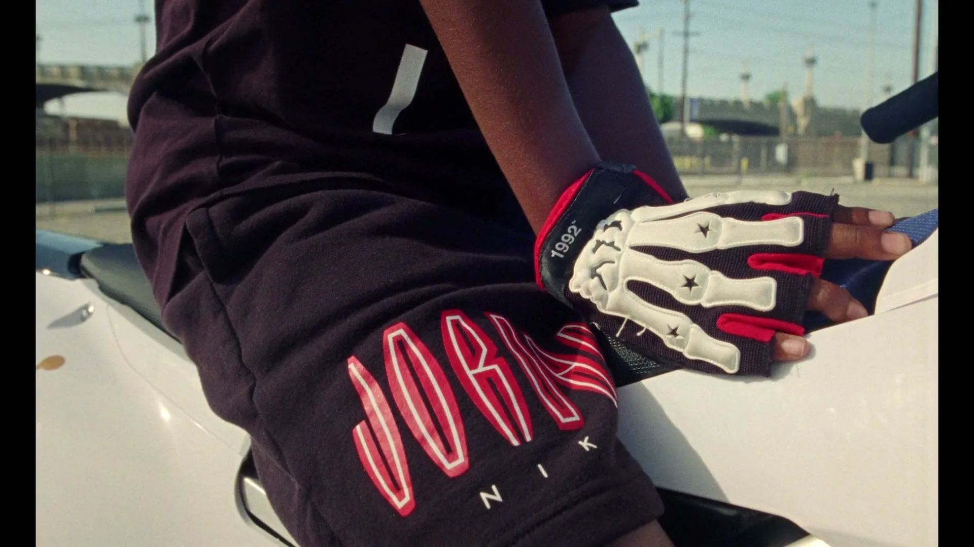 A person wearing black shorts with a red Jordan logo and a skeleton-hand glove sits on a white motorcycle outdoors.