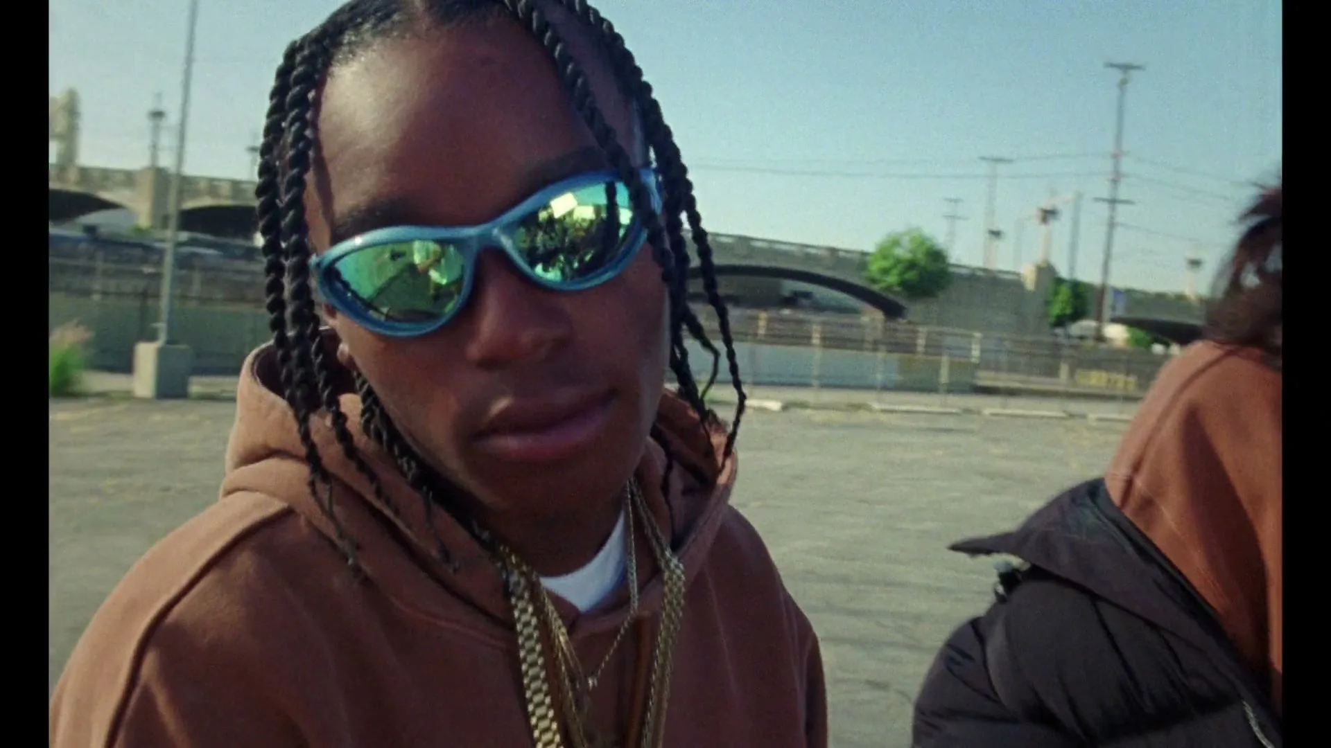 A young man with braided hair and blue reflective sunglasses wears a brown hoodie and gold chains, looking at the camera in an outdoor parking lot.