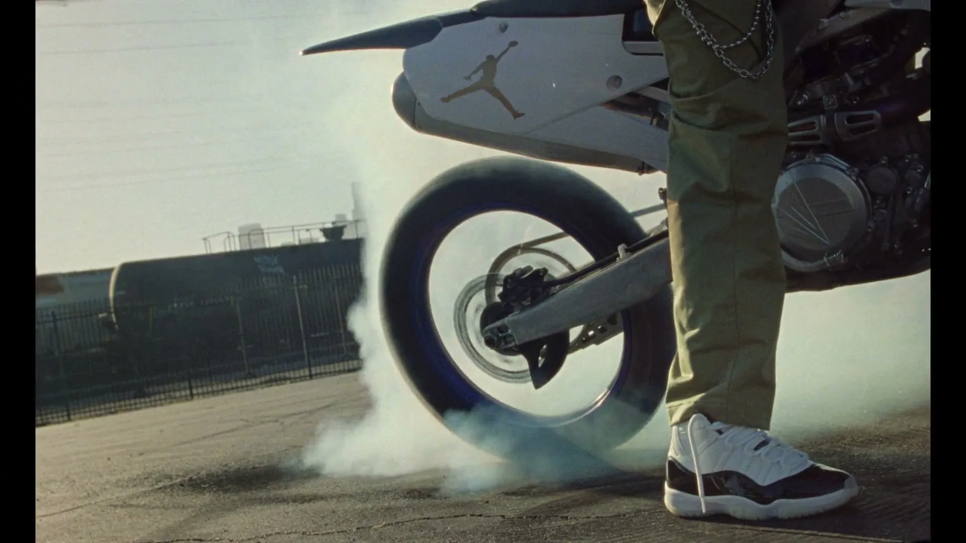 A person in cargo pants and Jordan sneakers stands next to a motorcycle doing a burnout, with smoke billowing from the rear tire on an asphalt surface.