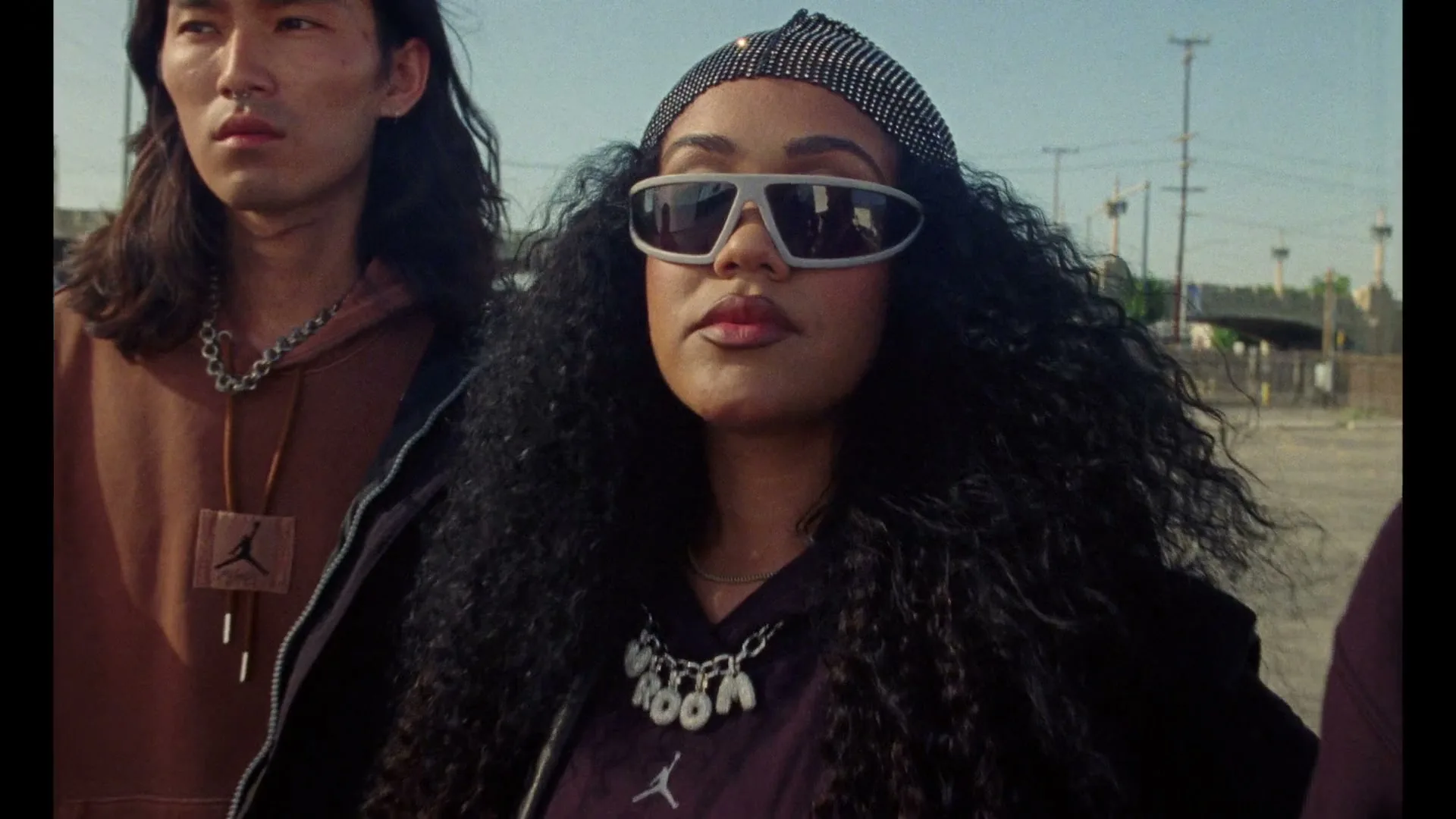 A diverse group of young adults, including a woman in sunglasses and a man with long hair, stand outdoors in an urban setting.