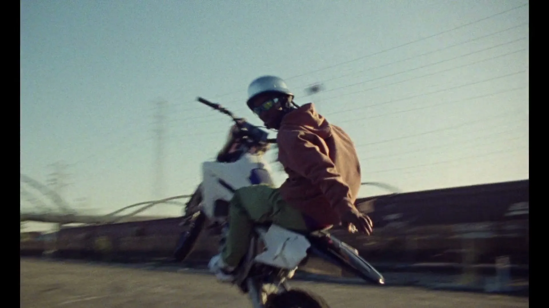 A person in a helmet and sunglasses rides a dirt bike on one wheel, performing a wheelie in an urban industrial area during the day.