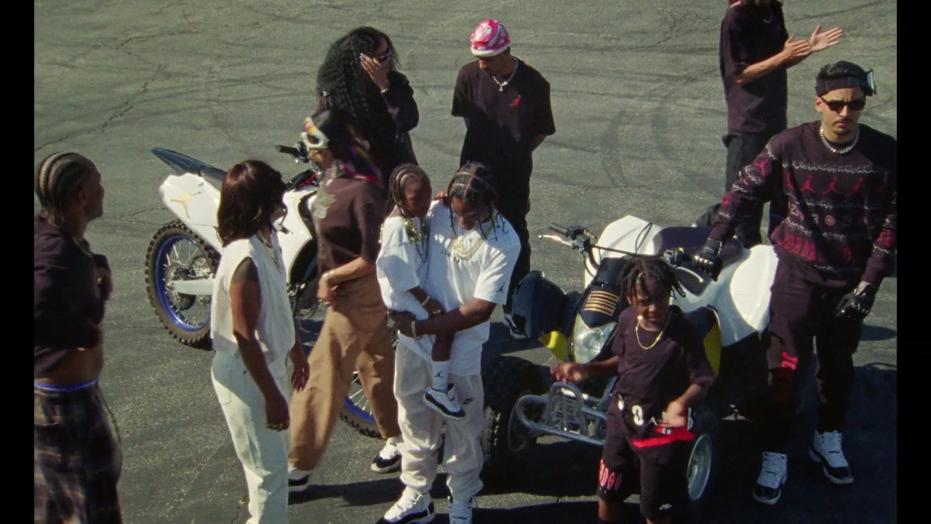 A diverse group of people, including adults and children, gather around dirt bikes and ATVs in a sunlit outdoor parking lot.