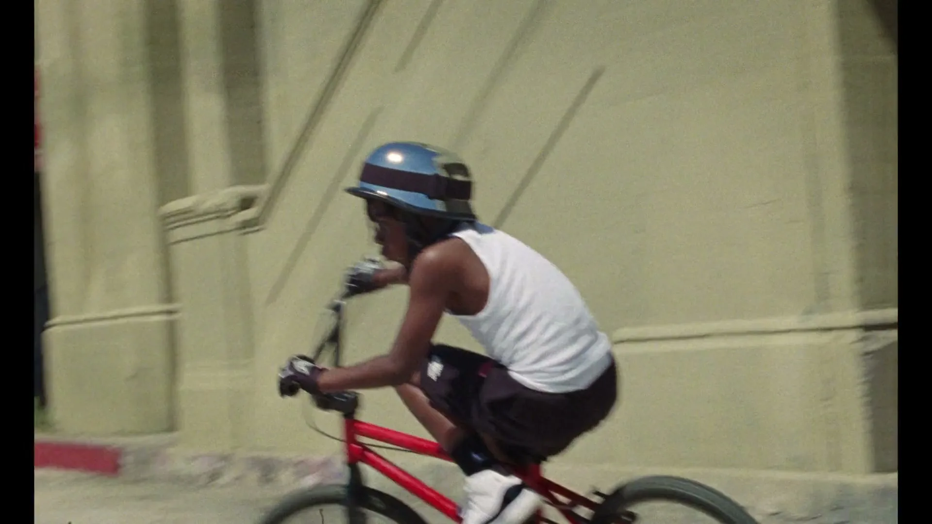 A young boy in a helmet and tank top rides a red BMX bike down a sunlit street, then sits next to it as a dirt bike speeds past.