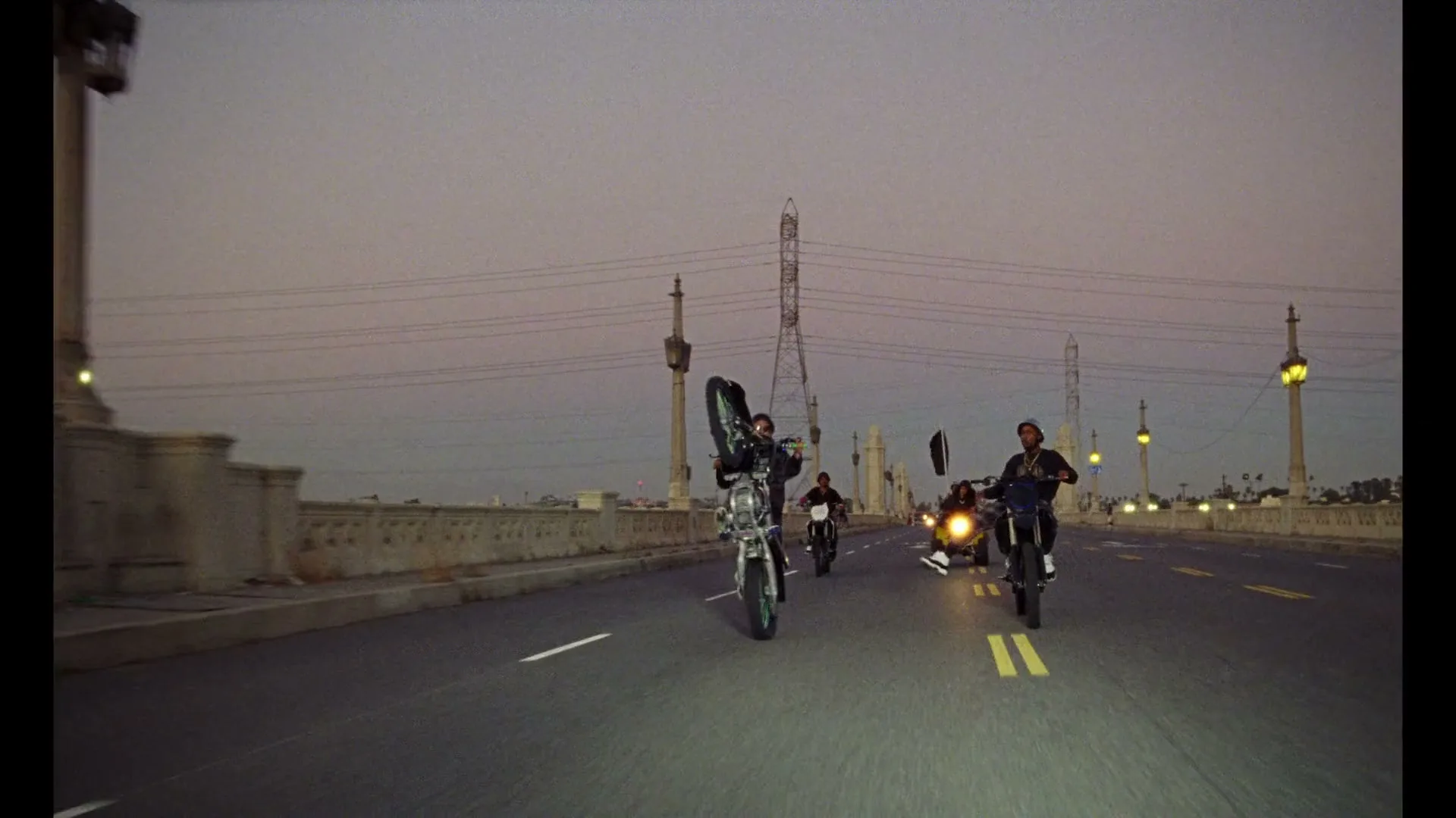 Three young men ride motorcycles on a bridge at dusk, performing wheelies and stunts, with city lights in the background.