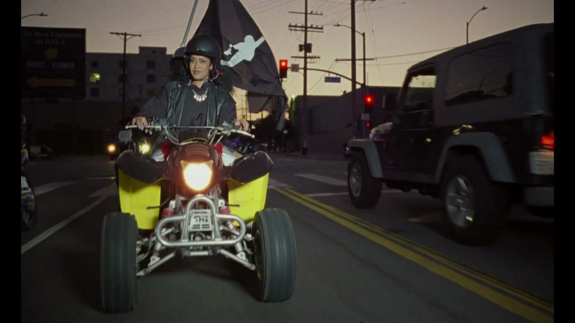 A woman in a black helmet and jacket rides a yellow ATV down a city street at dusk, holding a black flag with a white logo.