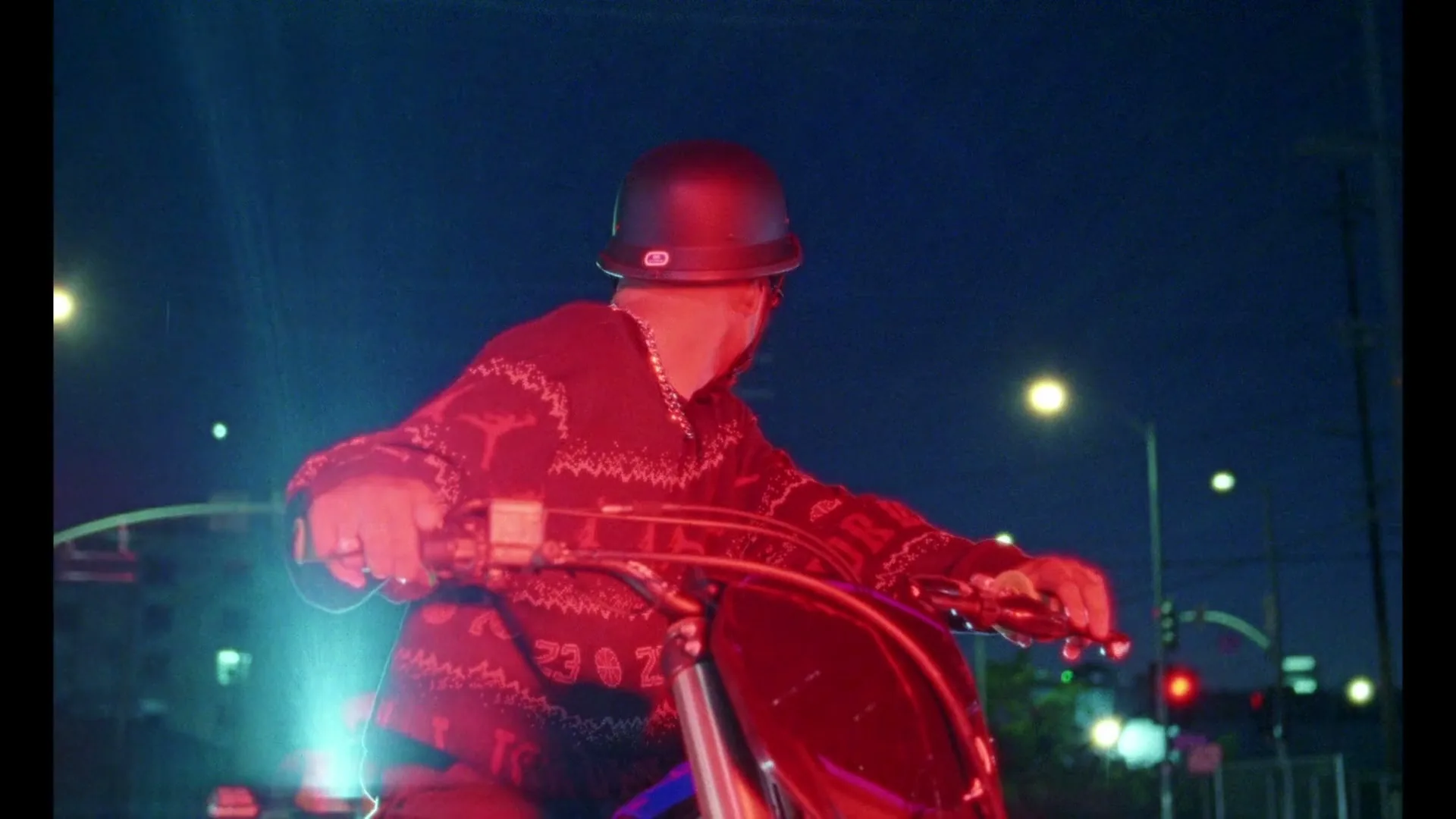A person wearing a black helmet and a red patterned sweater rides a motorcycle at night, with city lights in the background.