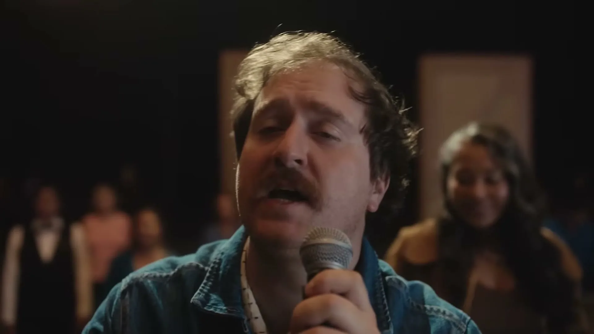 A man with a mustache sings into a microphone, eyes closed, with a blurred audience in the background.