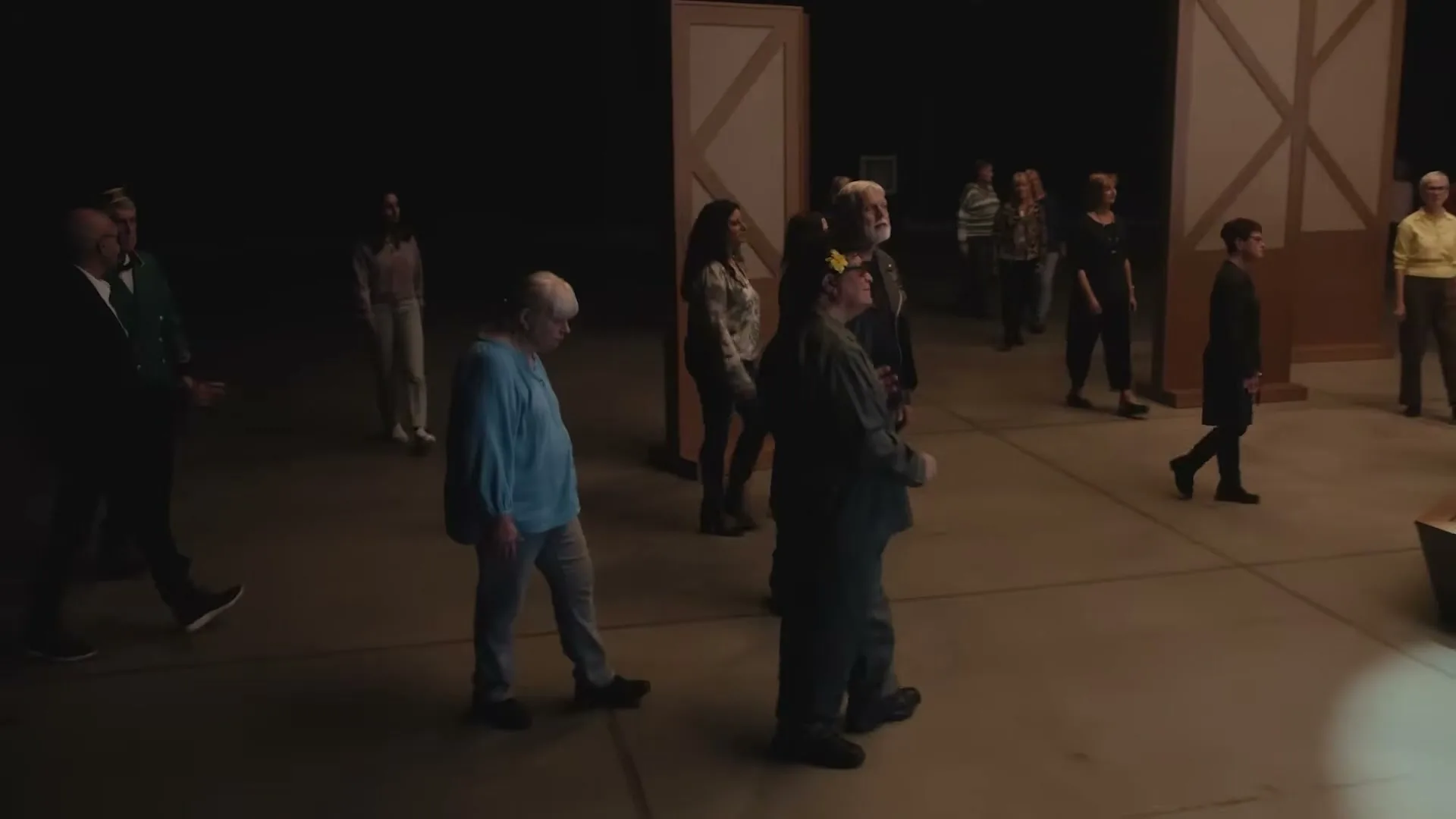 A diverse group of people, dressed in everyday clothes, walk slowly and aimlessly across a dimly lit stage, appearing lost and disoriented.