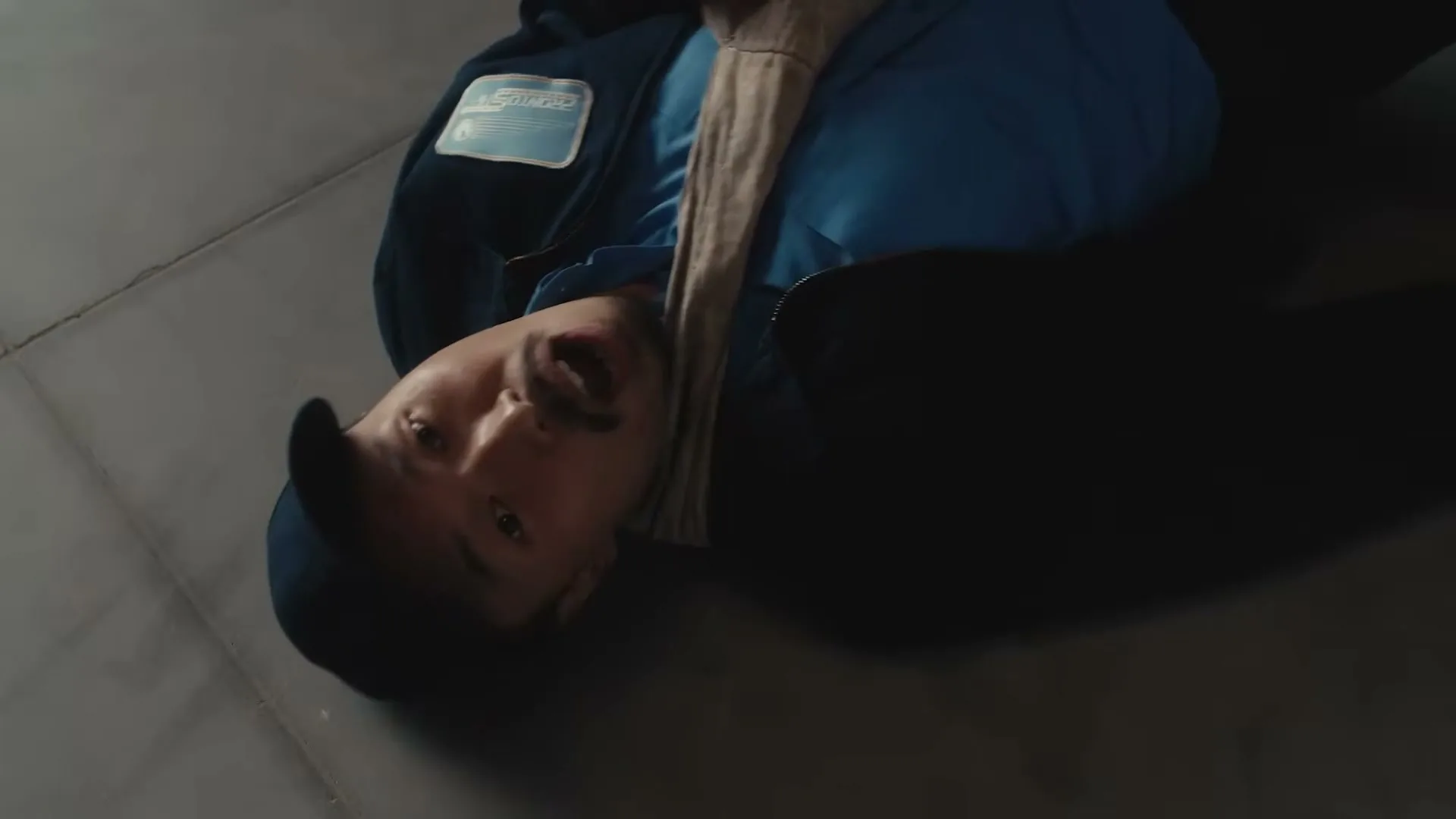 A man in his 30s, wearing a blue uniform and cap, lies on the floor looking up at the camera with a worried expression.