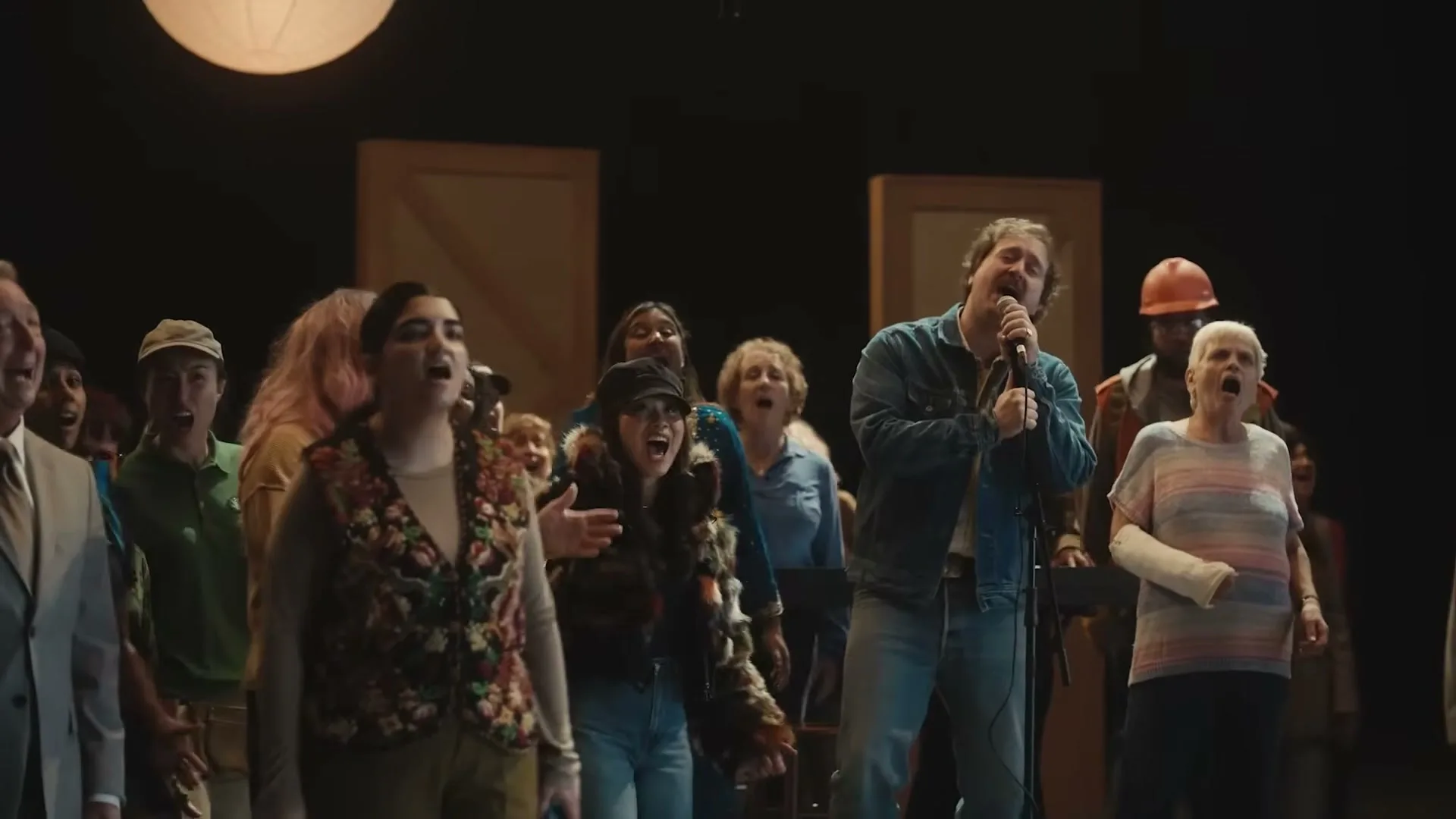 A diverse group of people, including a man singing into a microphone, stand together on a stage, all looking up and singing with open mouths.