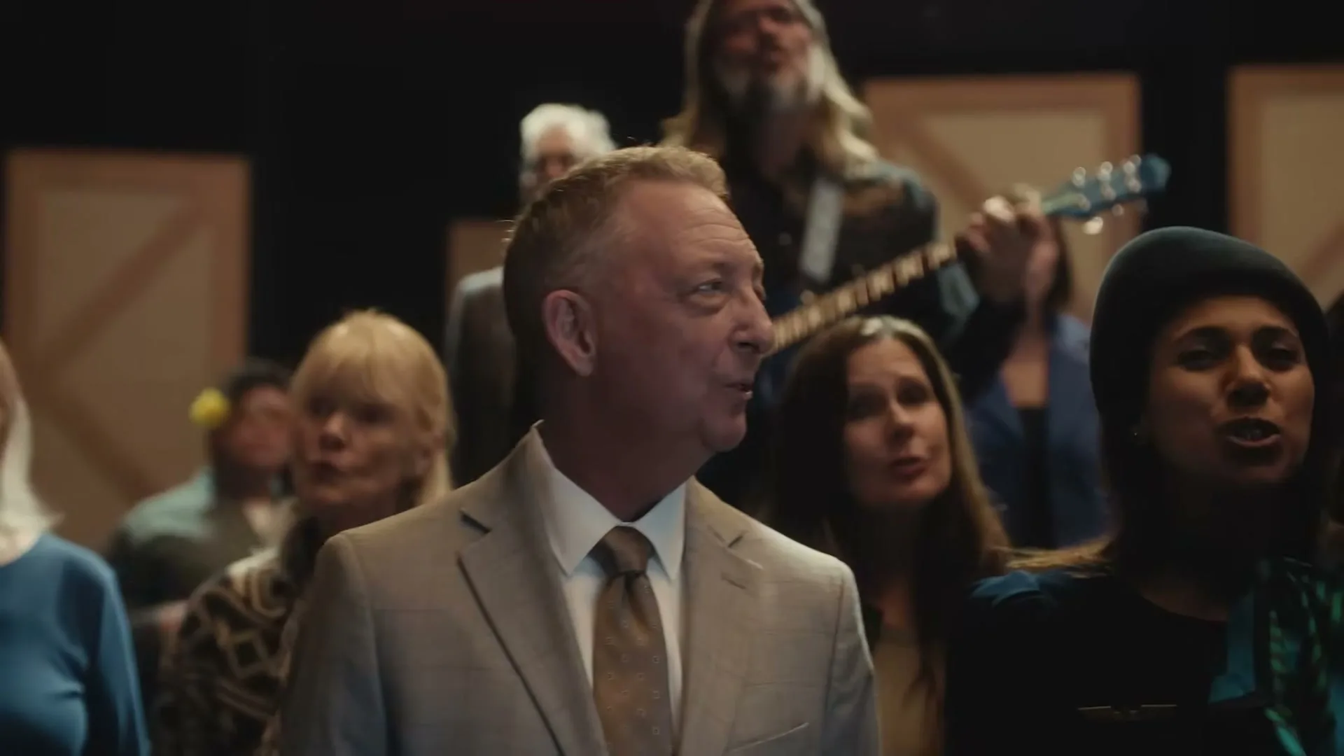 A middle-aged man in a suit sings with a diverse group of people in a choir, looking around with a slightly concerned expression in a dimly lit studio.