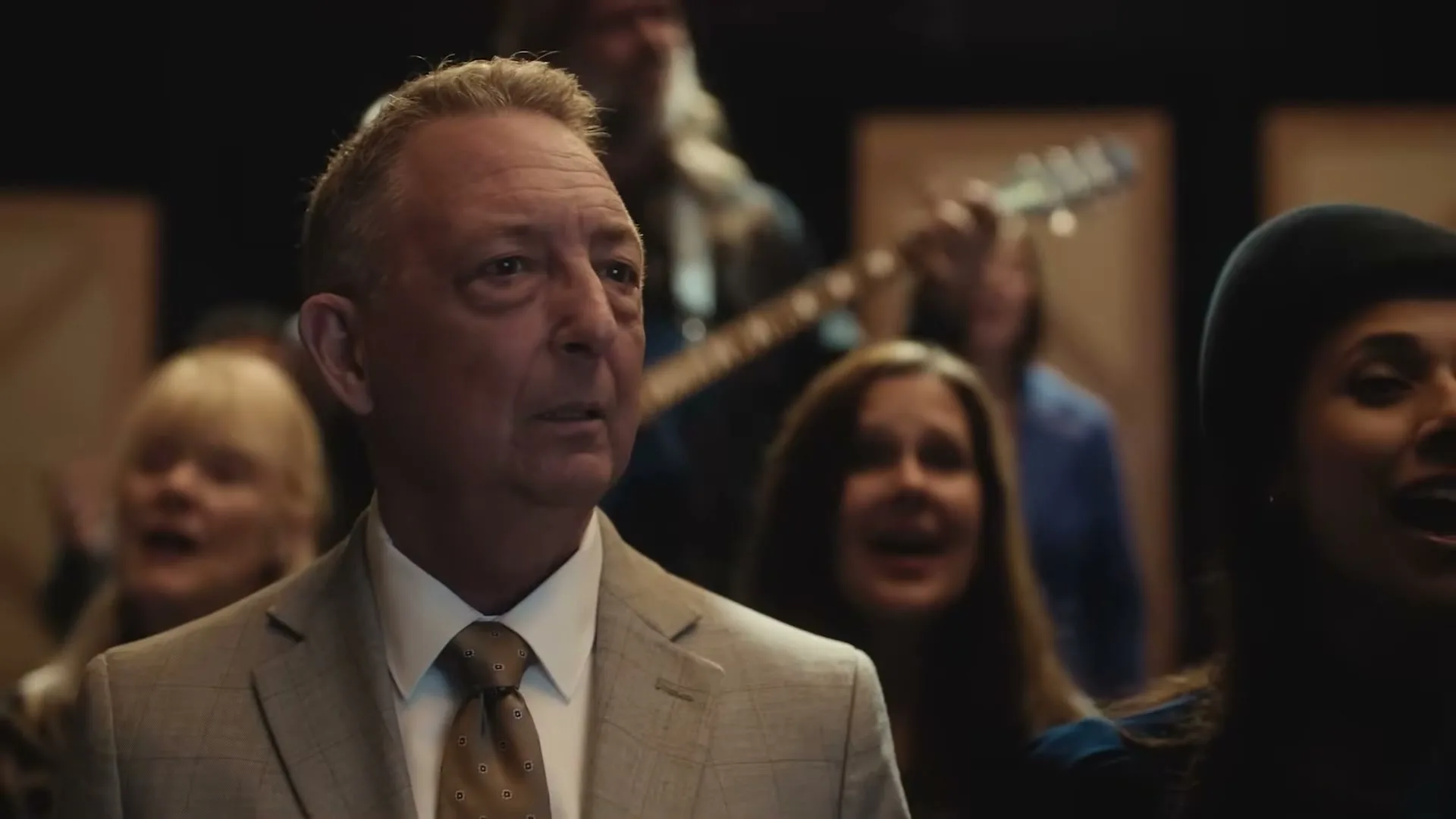 A middle-aged man in a suit looks concerned while surrounded by other people singing in what appears to be a church or concert setting.