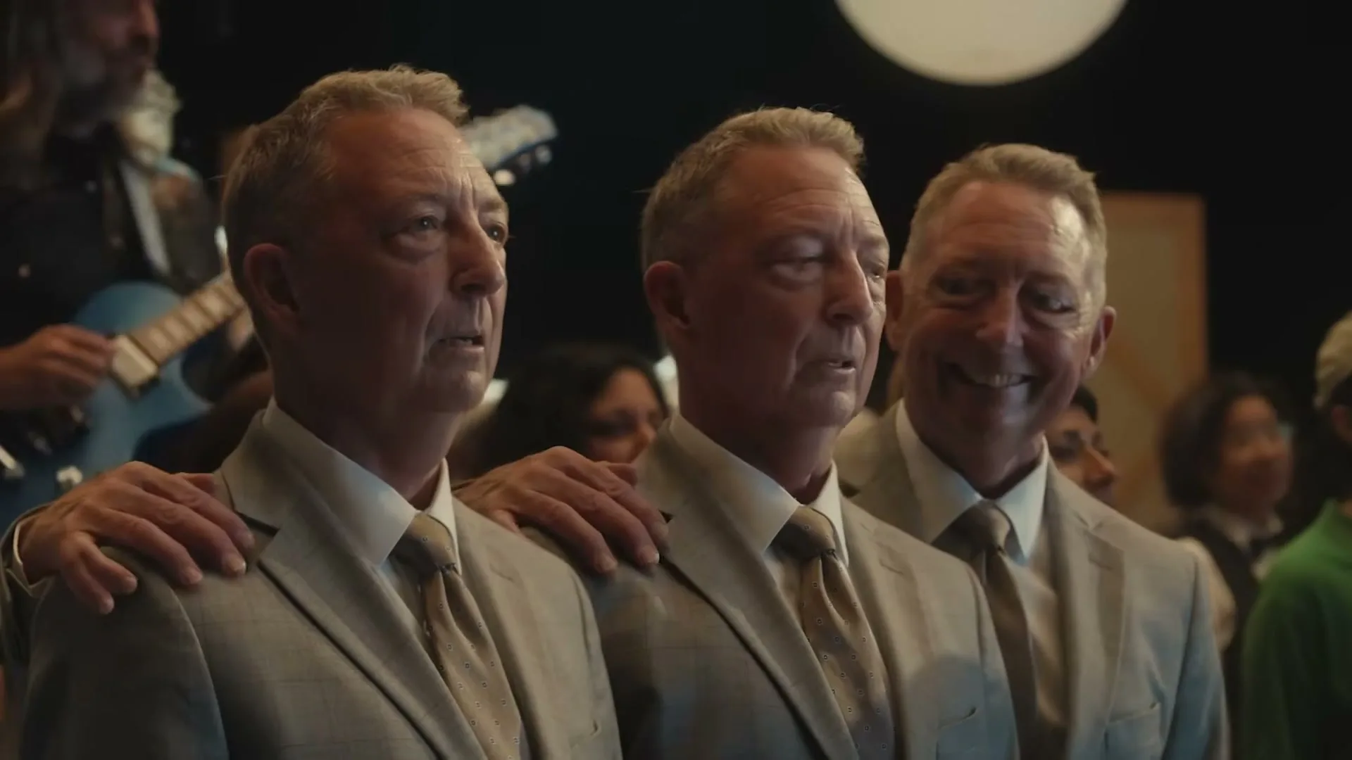 Three identical middle-aged men in suits stand side-by-side, smiling and looking forward, with a crowd and a guitarist in the background.