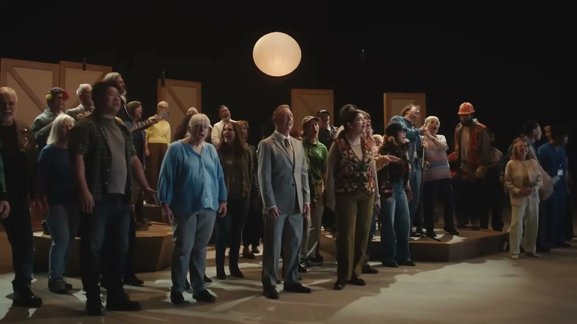 A diverse group of people, dressed in various casual and formal attire, stand together on a stage with a large paper lantern overhead, looking up and singing.