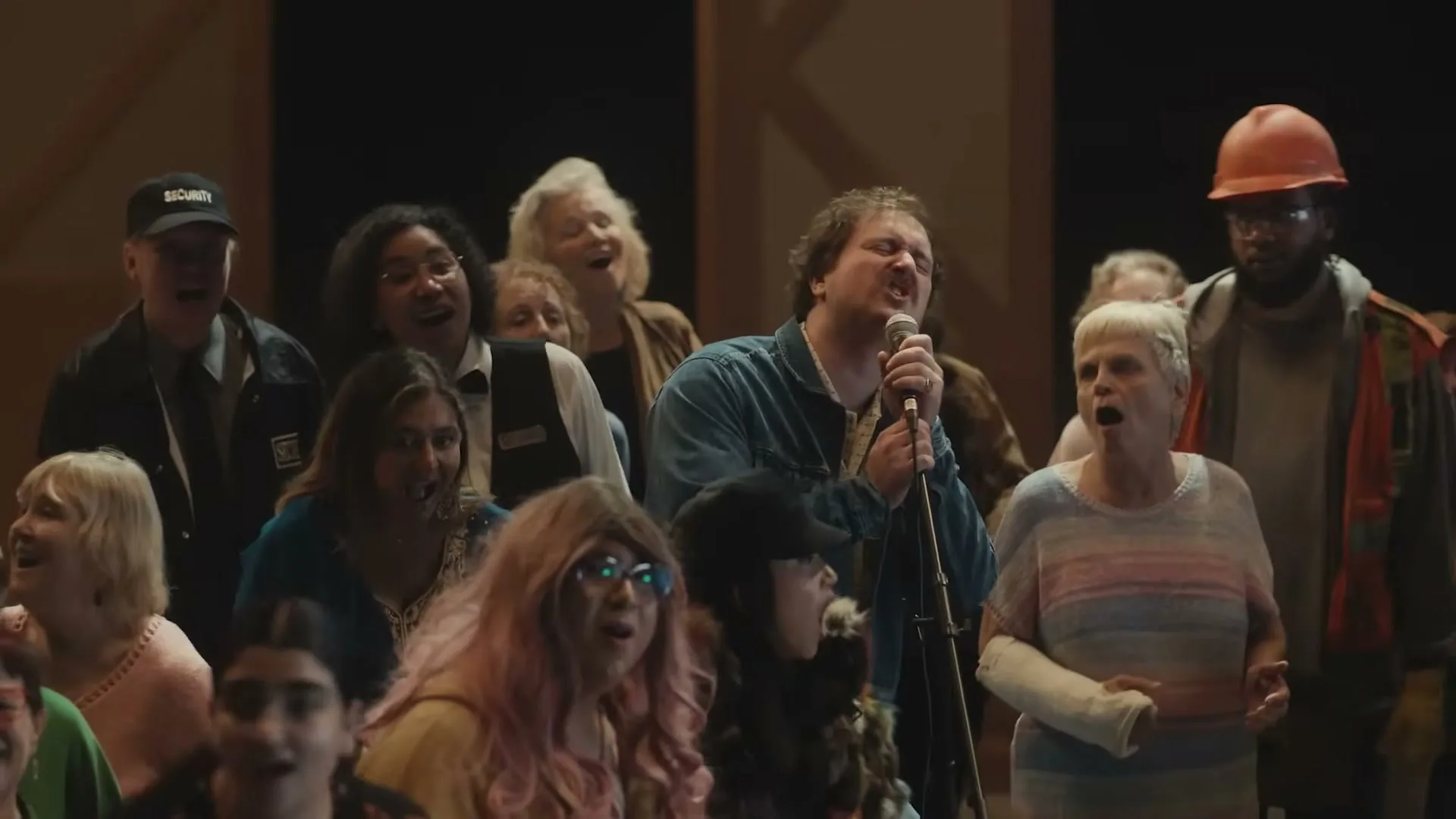 A diverse group of people, including a man singing into a microphone, are gathered together, all singing with open mouths in a dimly lit interior space.