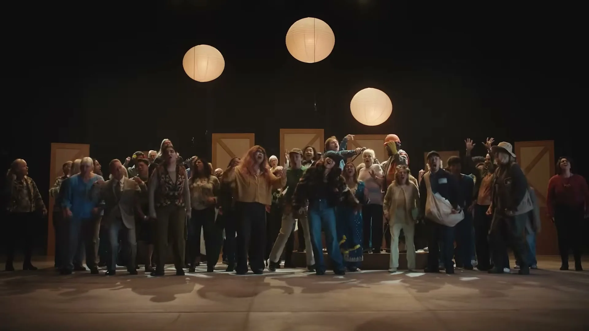 A diverse group of people, dressed in various casual outfits, stand on a stage with three paper lanterns hanging above them, looking concerned.