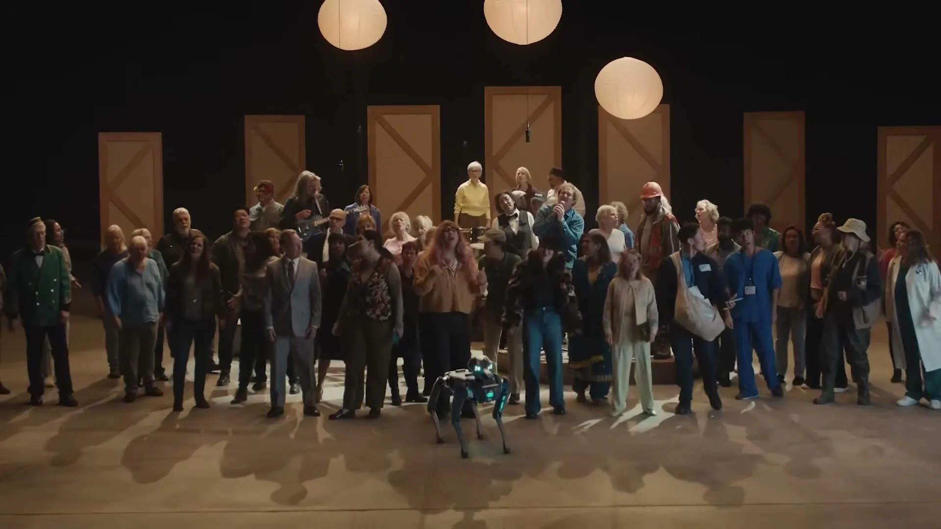 A diverse group of people, including a robot dog, stand on a stage with three paper lanterns and wooden panels in the background.