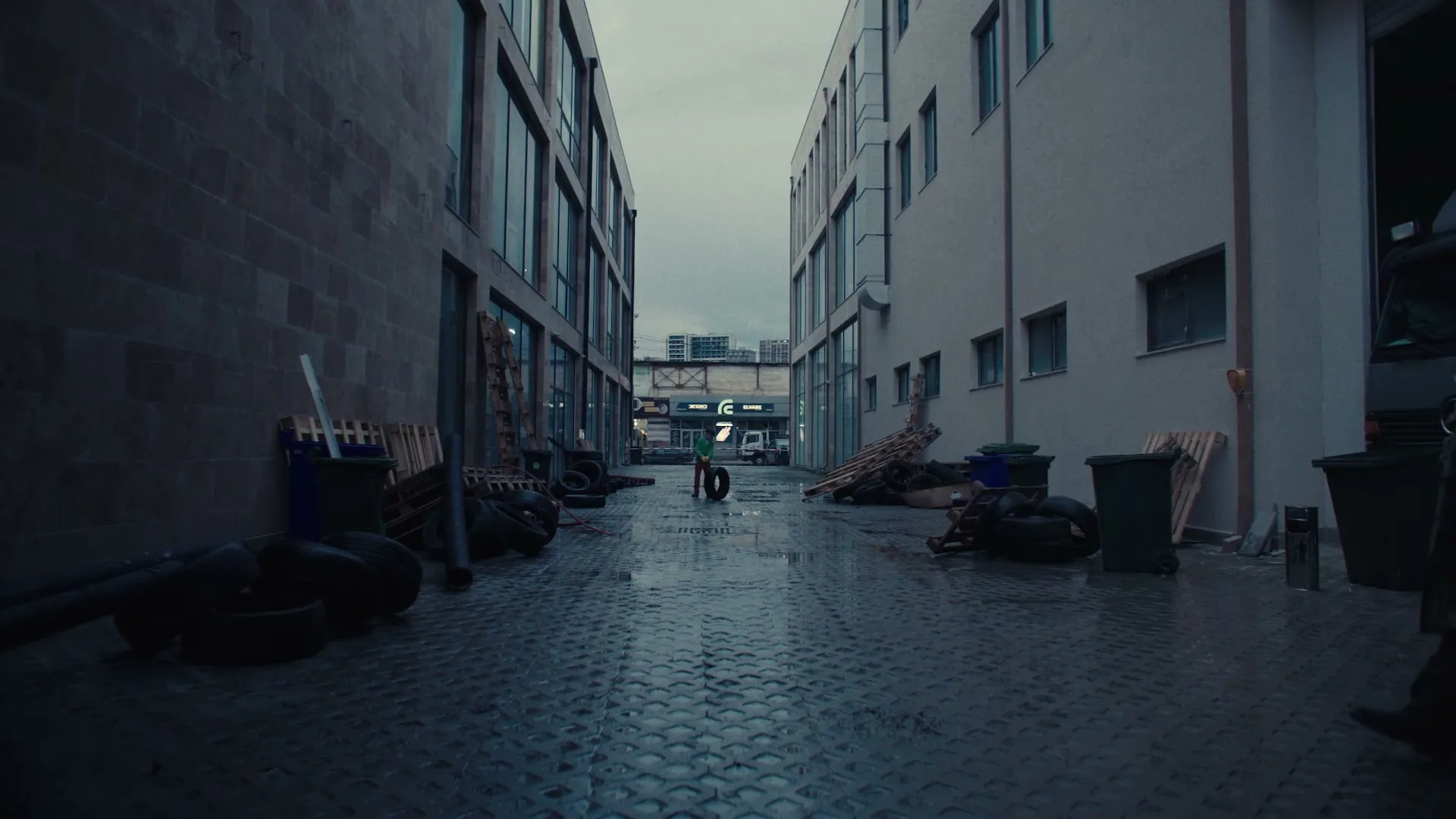 A wide shot of a dark, wet alleyway between two buildings, with a person in the distance rolling a tire and another person walking into frame on the right.