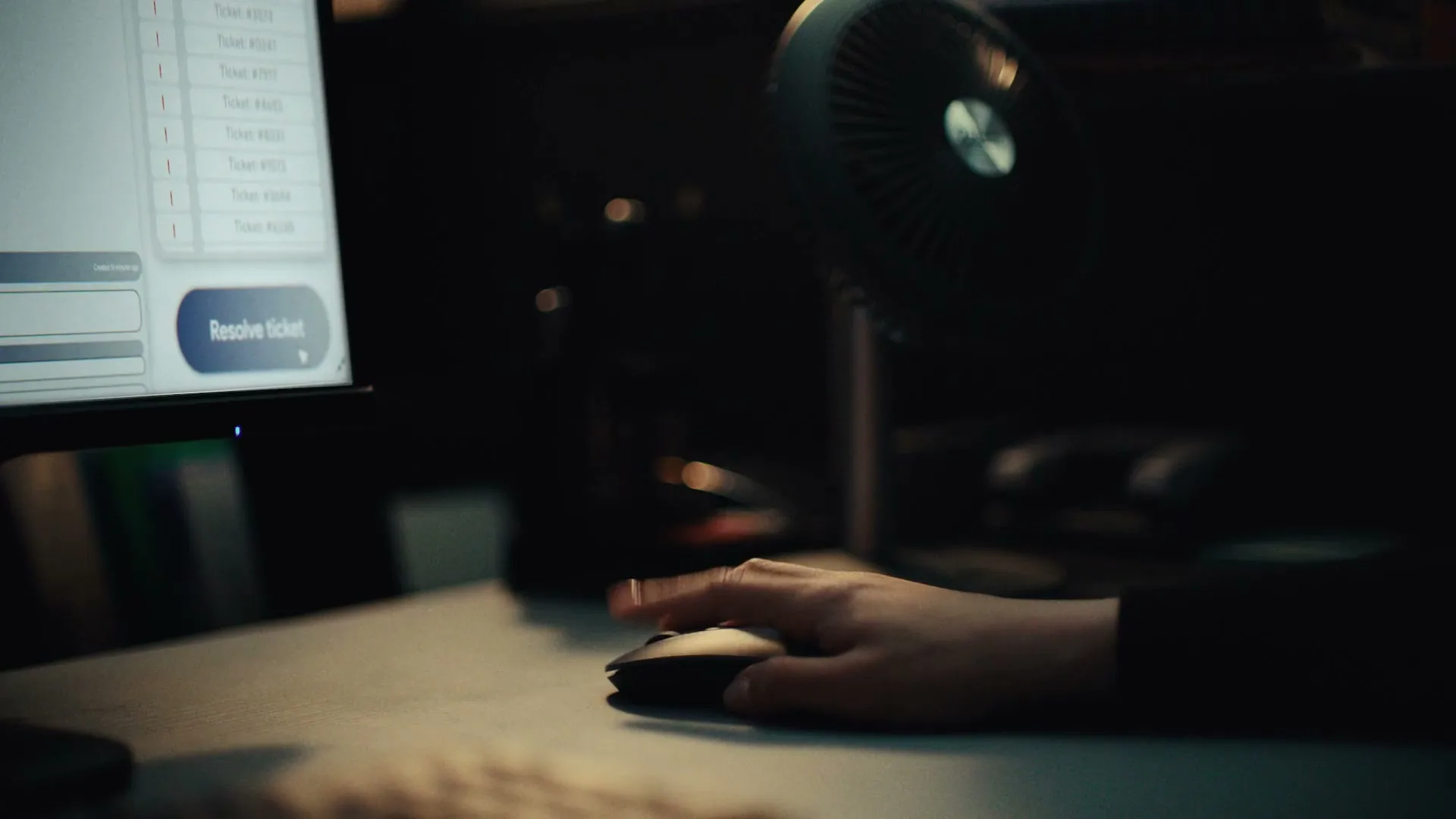 A person's hand clicks a computer mouse, with a monitor displaying a "Resolve Ticket" button and a small fan visible in a dimly lit office.
