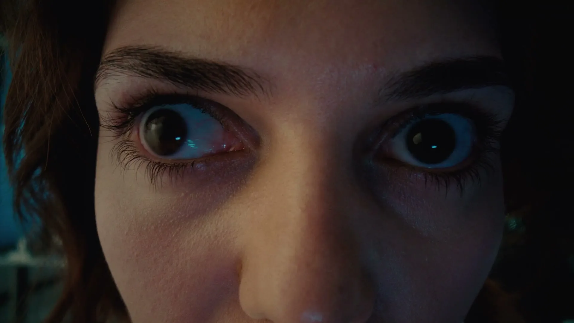 A woman's wide, bloodshot eyes stare intensely, reflecting a blue light, as she looks directly into the camera in a dark room.