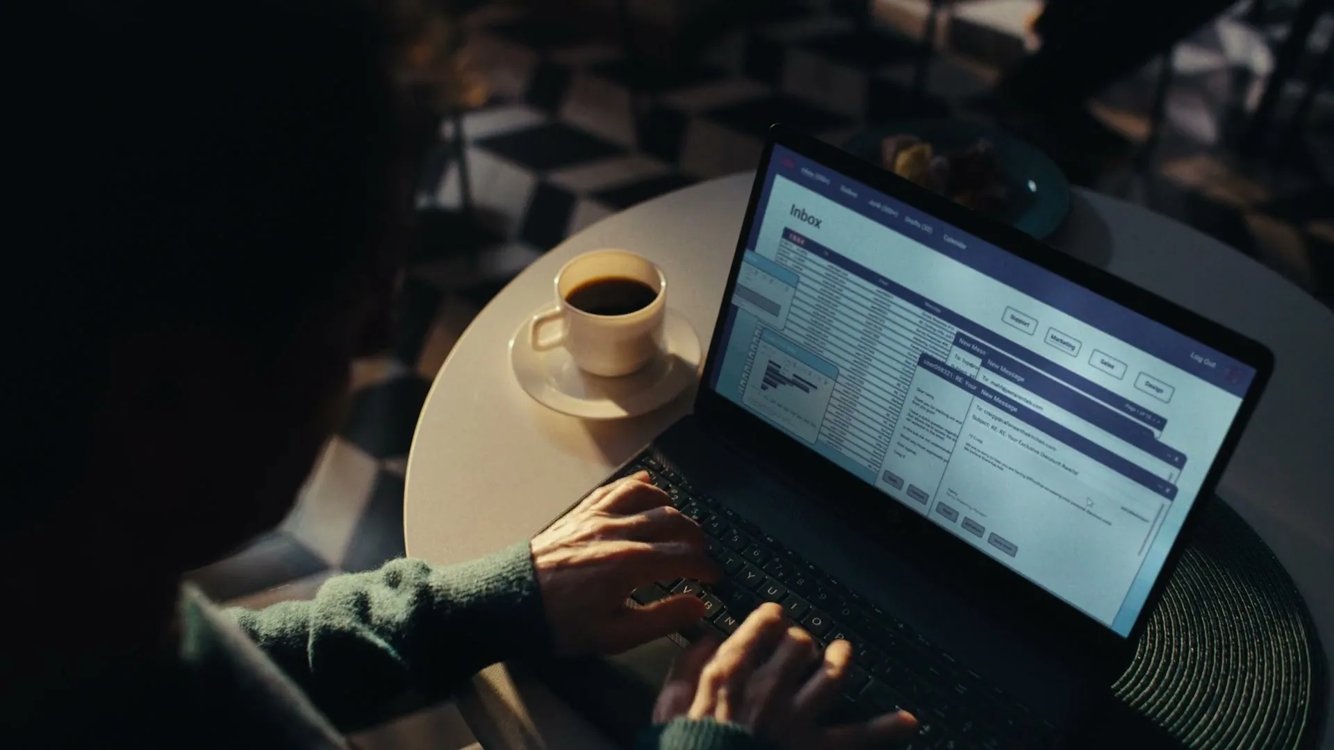 A person with dark hair and a green sweater types on a laptop, displaying an email inbox, while a cup of coffee sits on a white table in a dimly lit room.