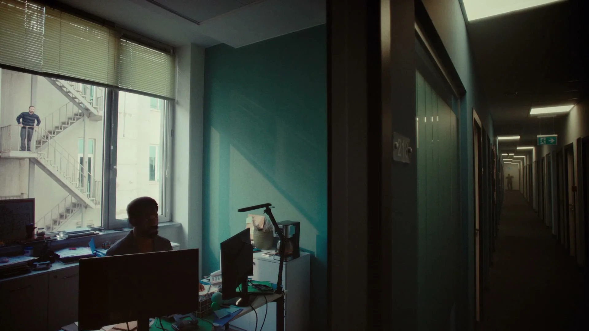 A man sits at a desk in an office, looking towards a long, dark hallway, while another man stands on an exterior fire escape.