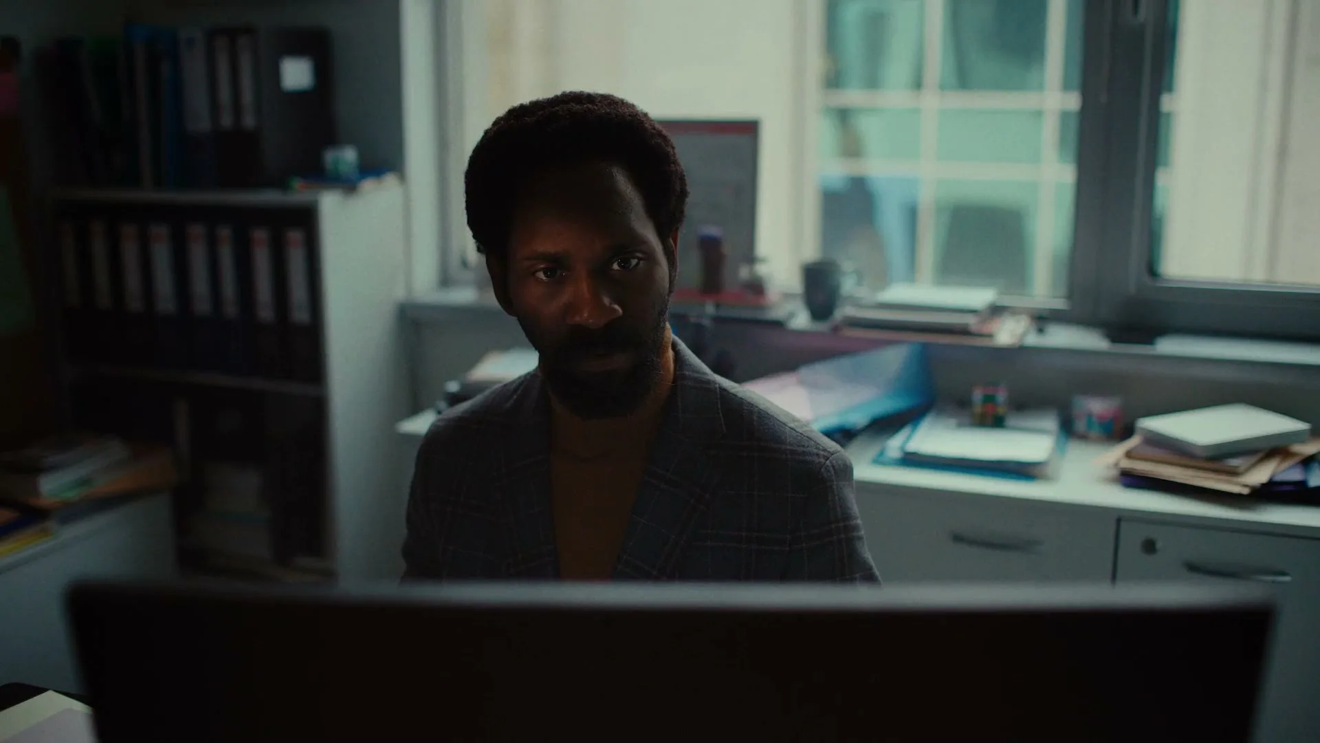 A Black man with a beard, wearing a plaid jacket and orange shirt, looks intently at a computer screen in a messy office.