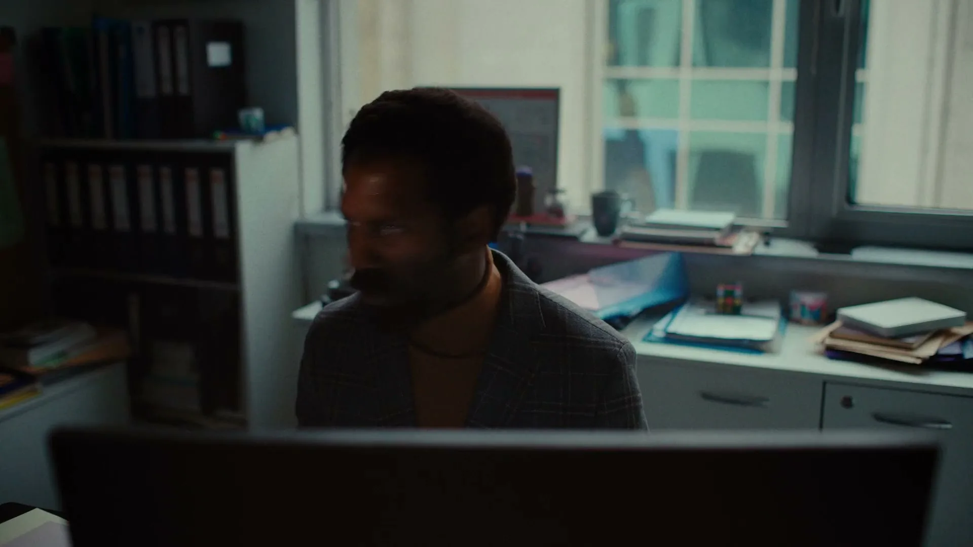 A man with dark skin and curly hair, wearing a plaid jacket, sits at a desk in an office, looking to the side.