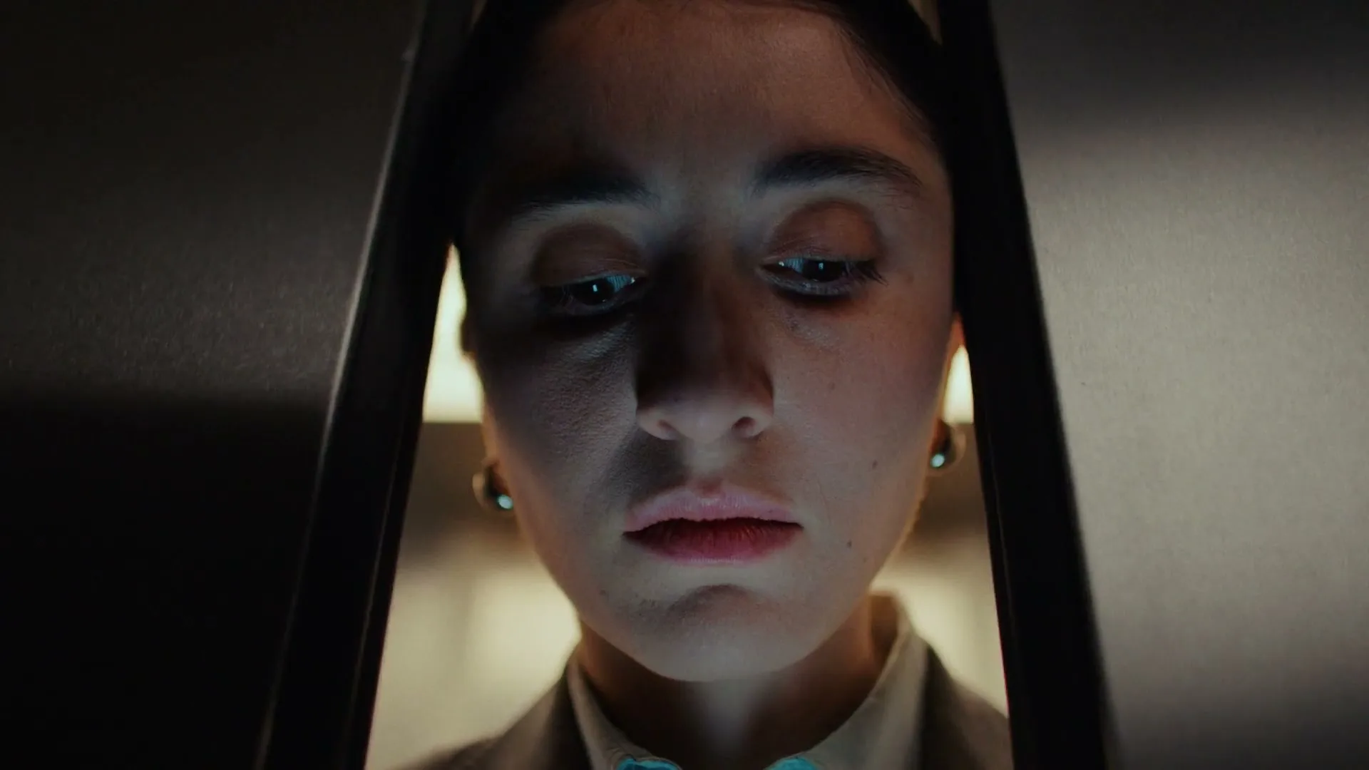 A young woman with dark hair, wearing a business suit, looks down intently at something off-screen, illuminated by a blue glow in a dark elevator.