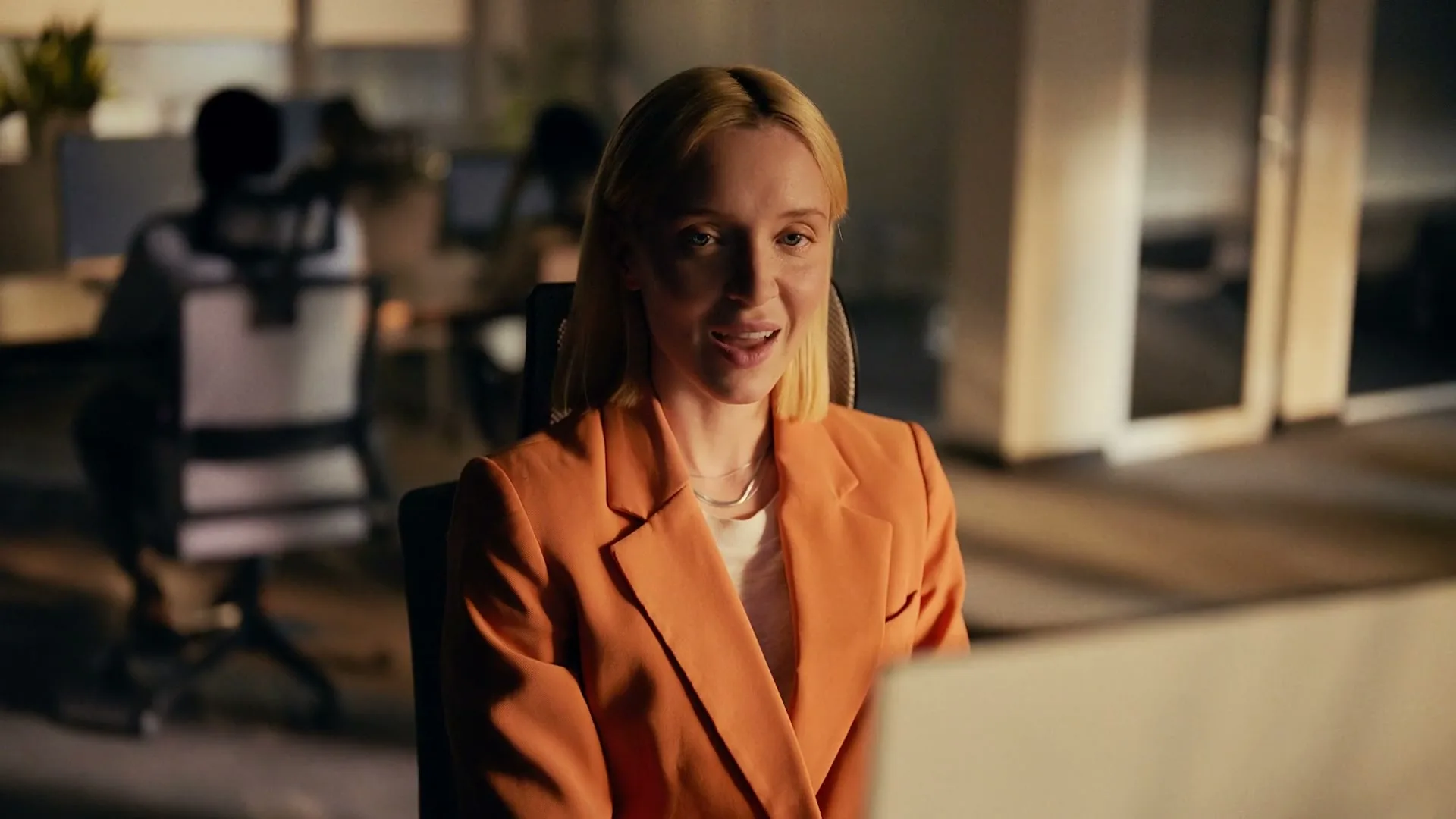 A blonde woman in an orange blazer looks up from her computer and smiles, while other colleagues work in the background of a modern office.