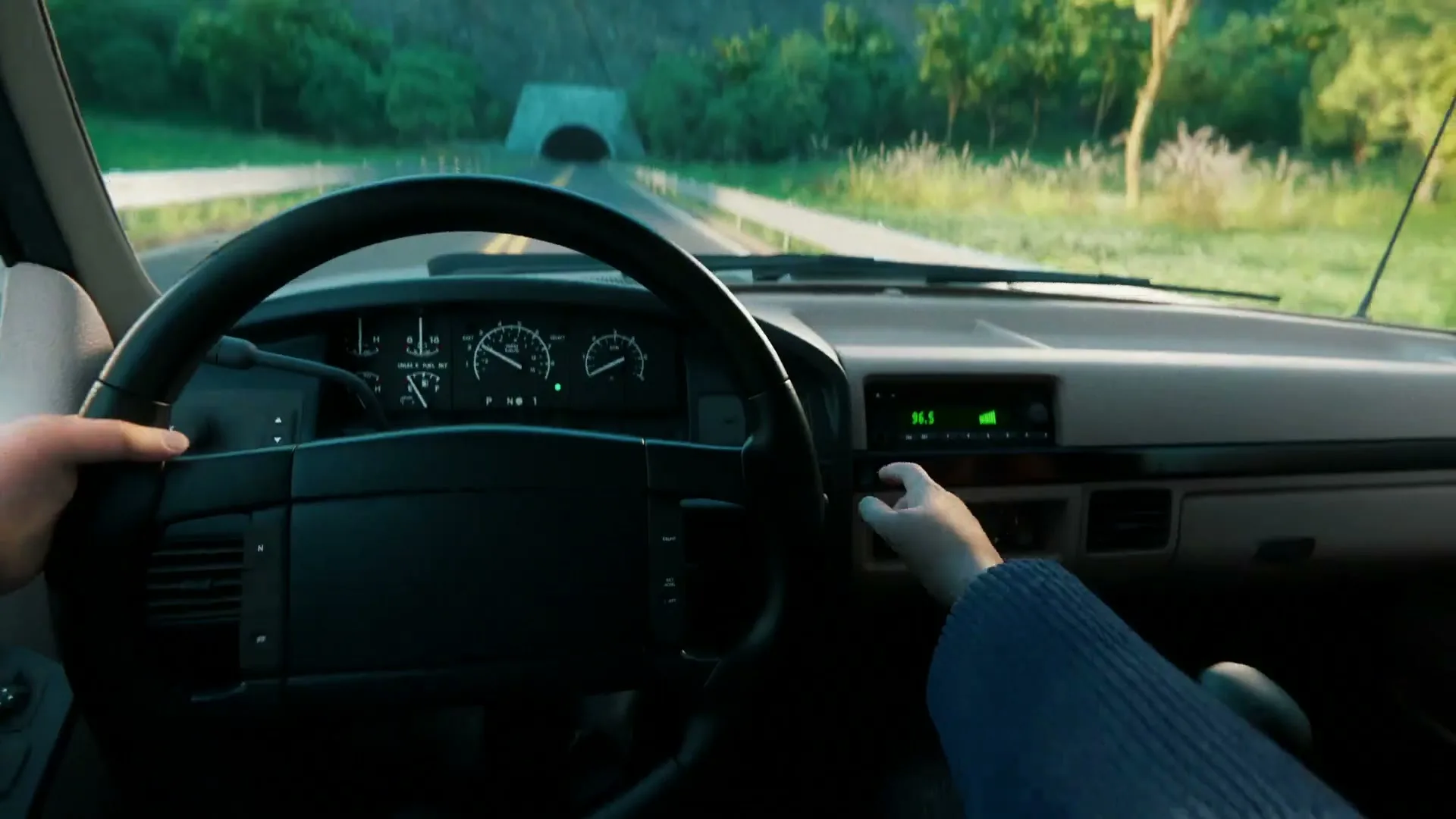 A person drives a car on a scenic road towards a tunnel, with their hands on the steering wheel and one hand adjusting the radio.