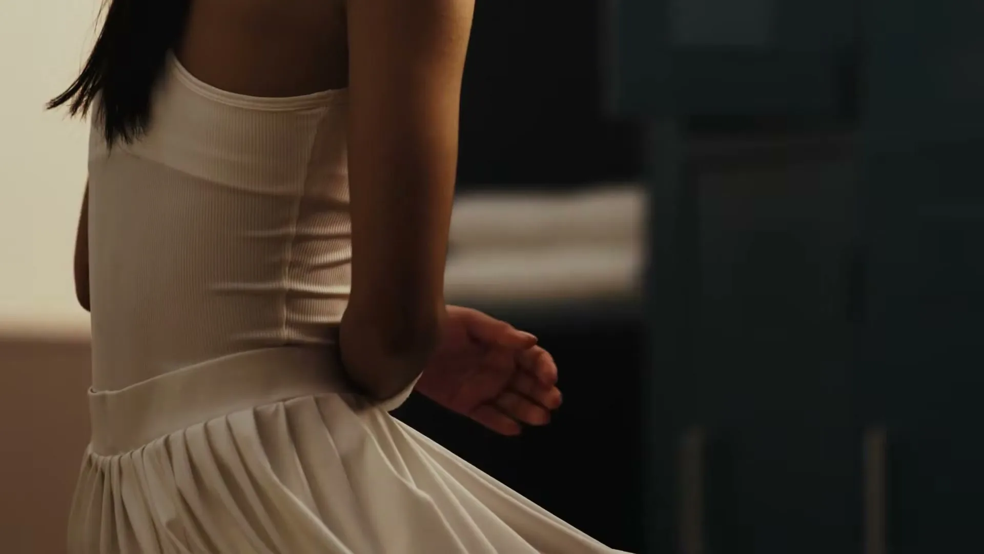 A young woman in a white dress sits, her hands clasped, in a dimly lit room, looking down with a pensive expression.