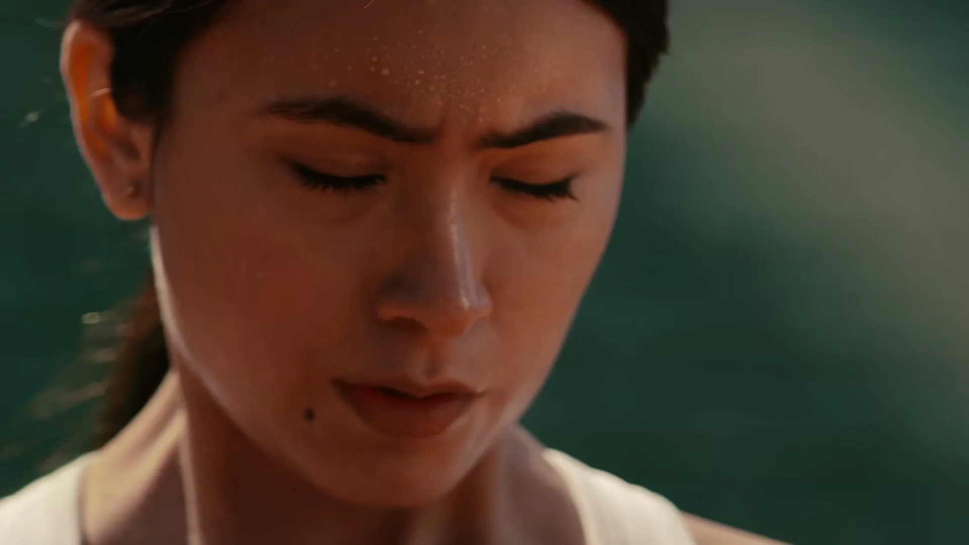 A young woman with sweat on her face, wearing a white tank top, looks down with a focused expression during an outdoor activity.