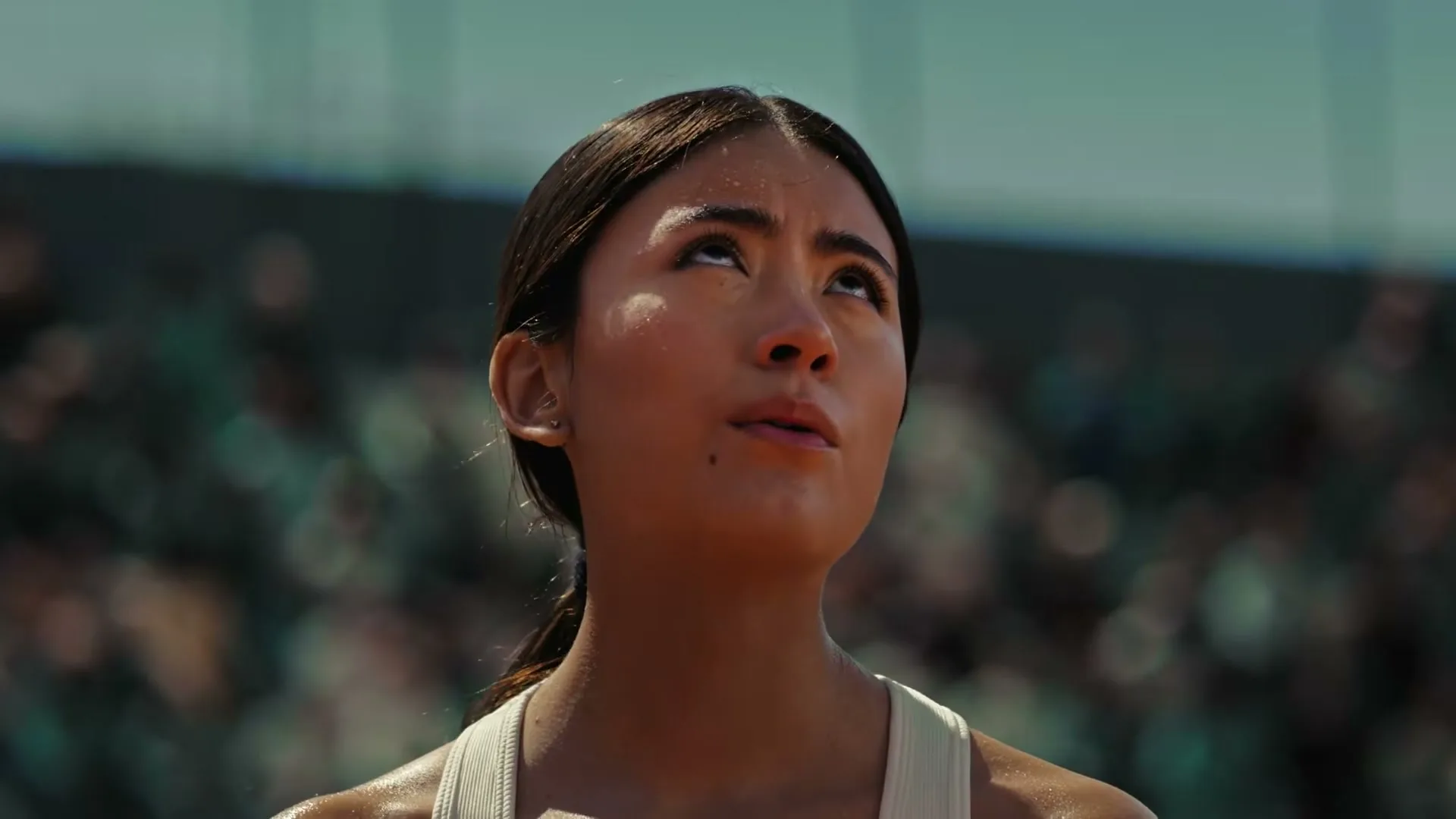 A young female tennis player, sweaty and focused, looks up with determination on a sunny outdoor court with blurred spectators in the background.