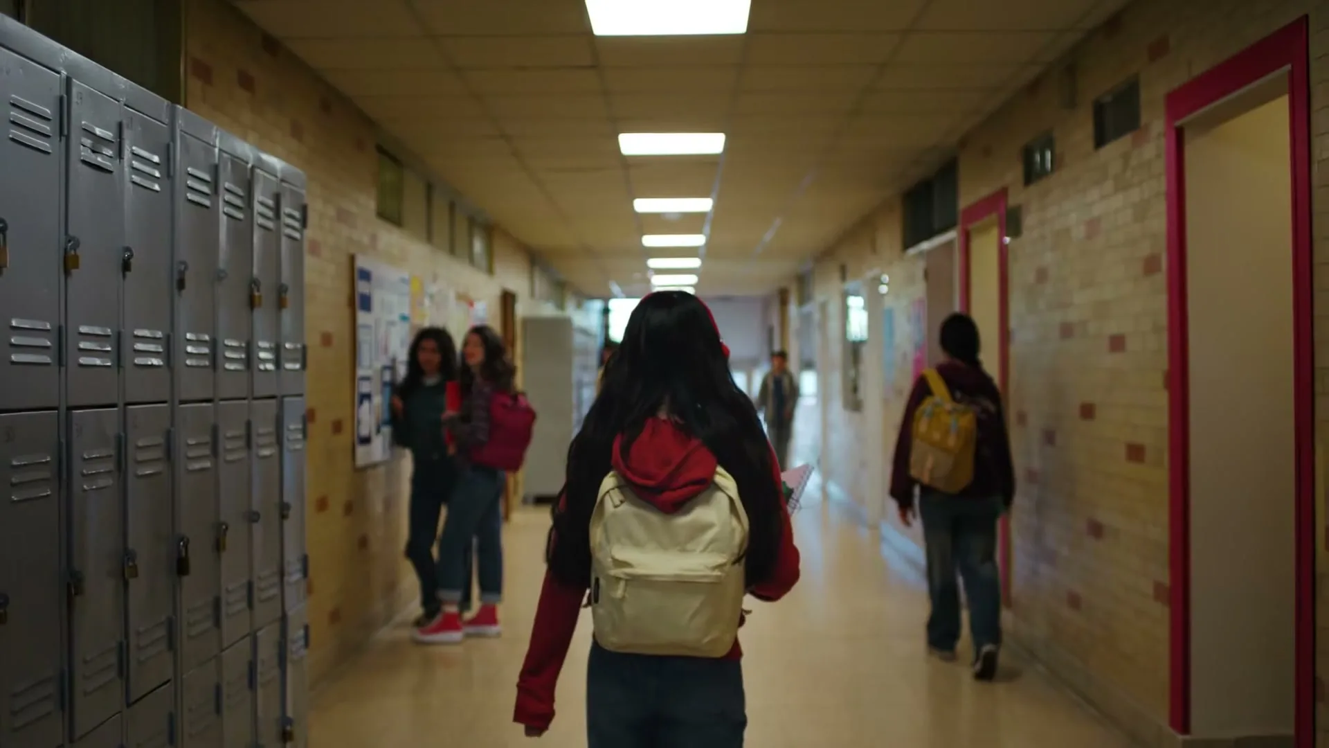 A young female student with long dark hair and a backpack walks down a school hallway, passing other students and lockers.