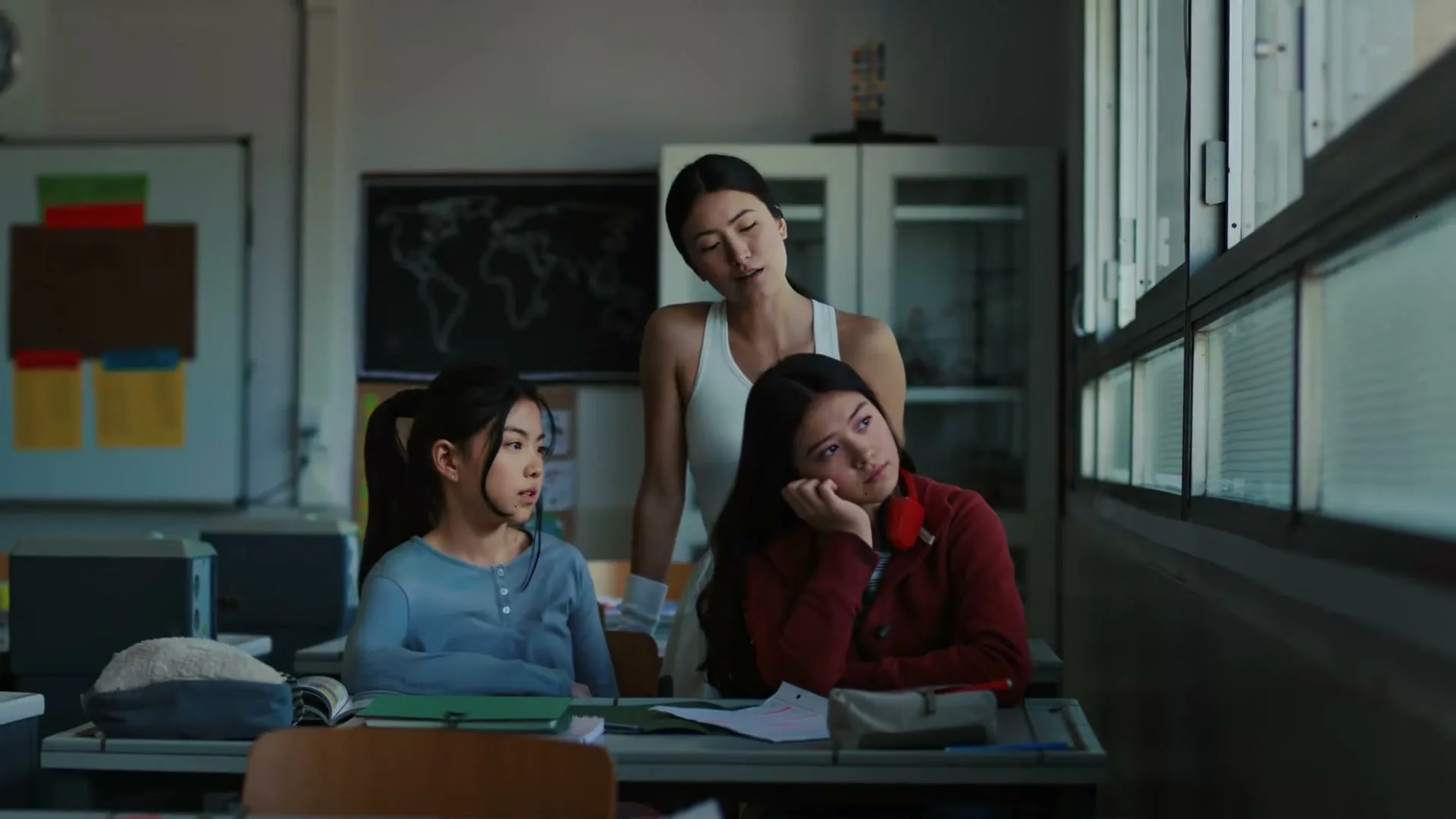 A young female teacher in a white tank top stands behind two female students in a classroom, placing a hand on one student's shoulder.