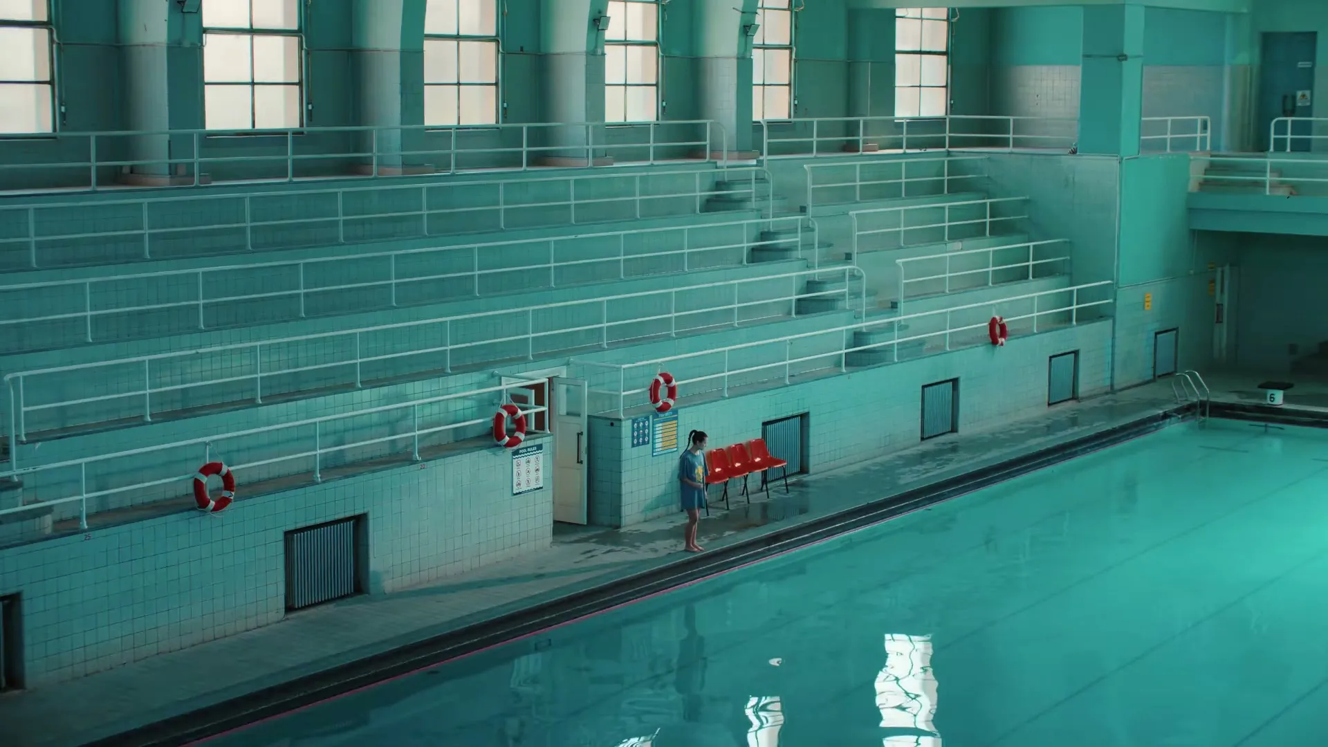 A young woman in a blue swimsuit stands by the edge of a large, empty indoor swimming pool with tiered seating.