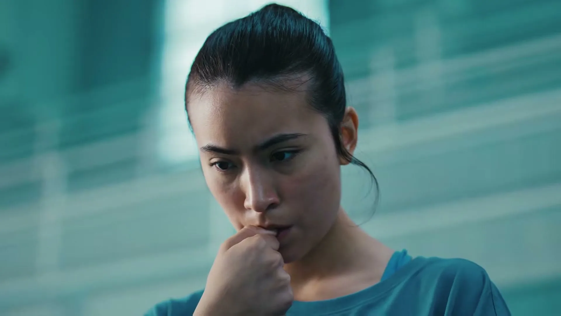 A young woman with dark hair pulled back, wearing a blue top, looks down thoughtfully, biting her finger in a modern, light-filled interior.