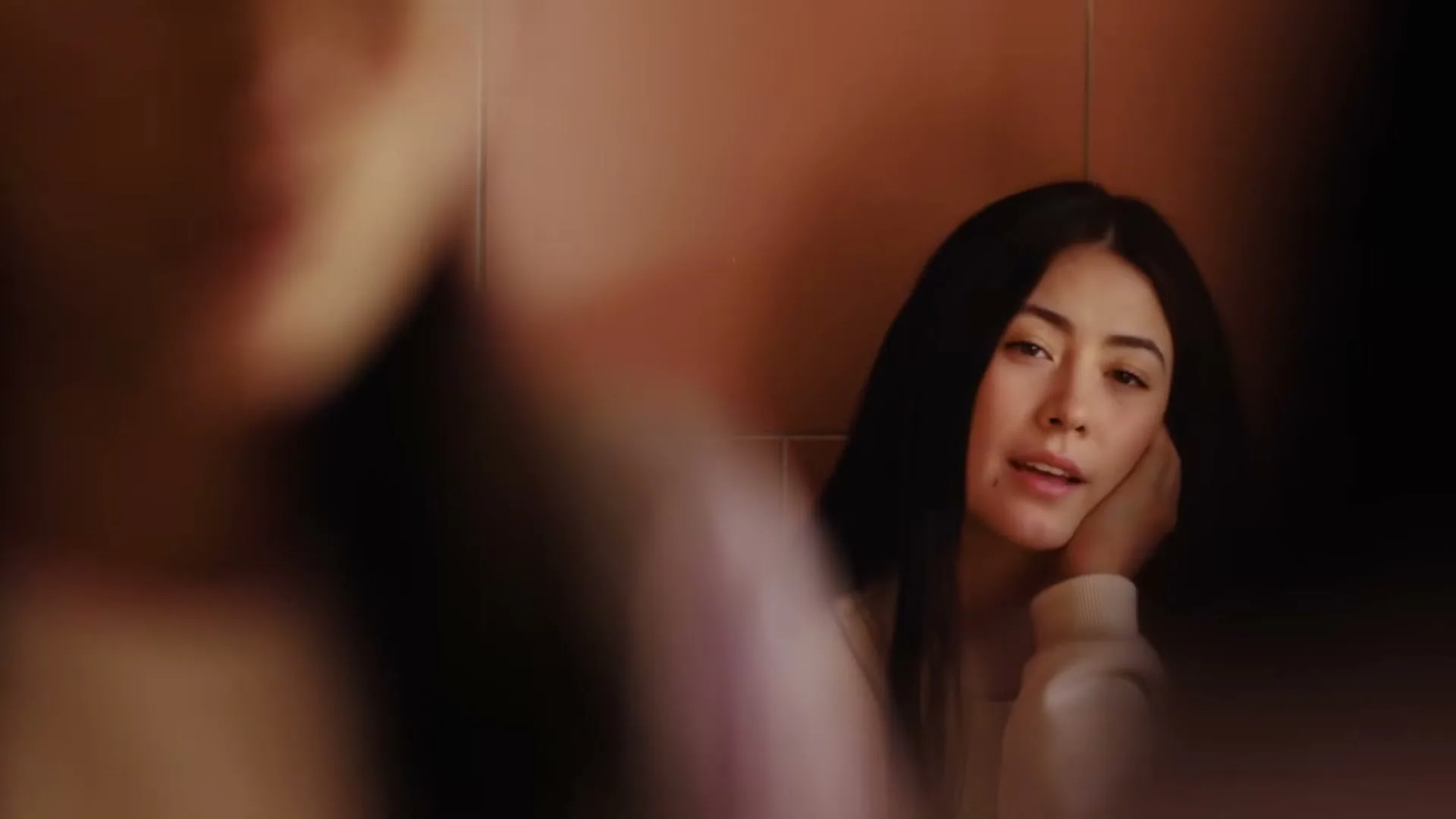 A young woman with long dark hair, wearing a white sweatshirt, looks thoughtfully into a mirror in a bathroom.