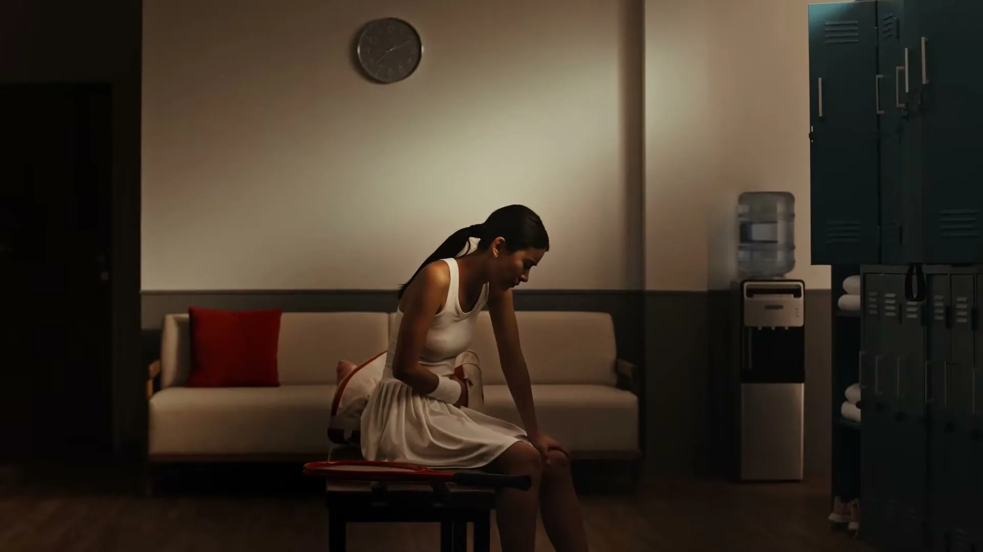 A young female tennis player sits on a bench in a locker room, clutching her stomach in discomfort.
