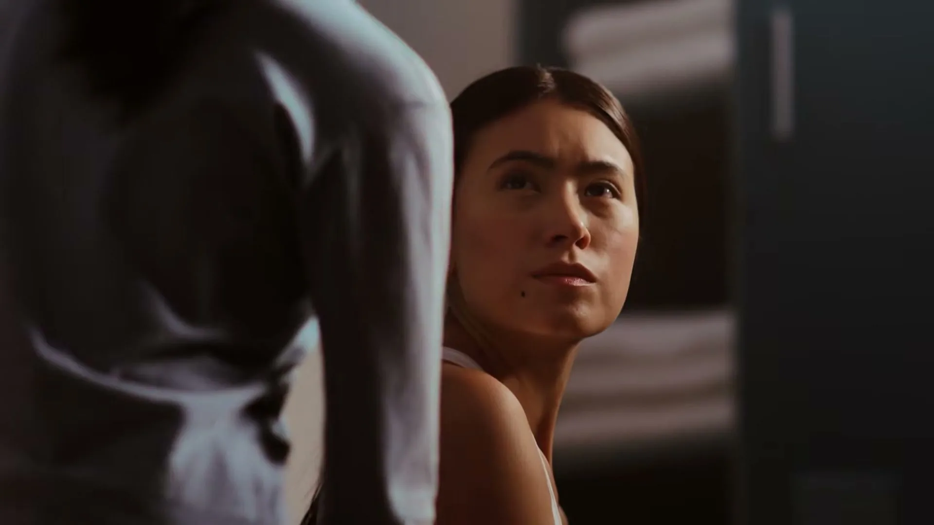 A young woman with dark hair looks up and to the right, while another person's back is visible in the foreground, in an interior setting.
