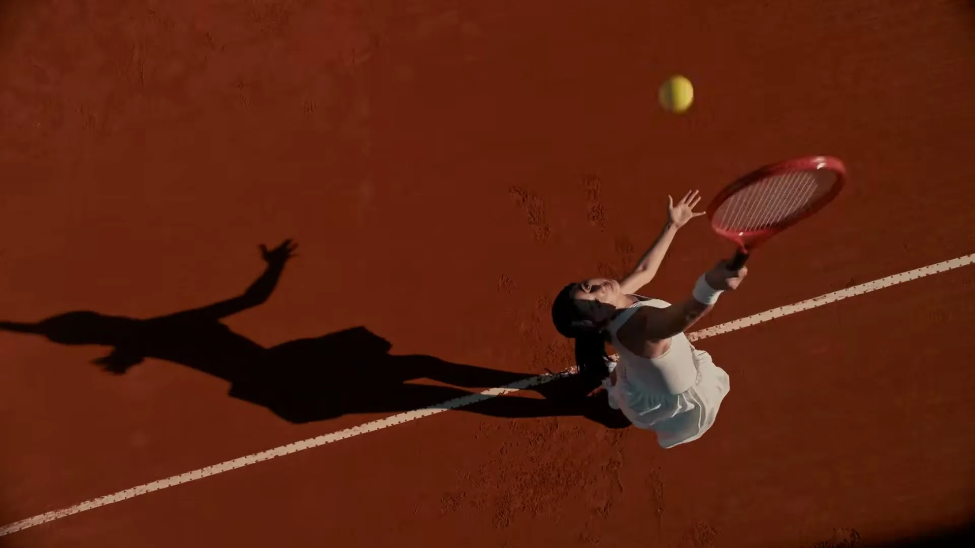 A female tennis player in a white dress serves a yellow tennis ball on a clay court, casting a long shadow.