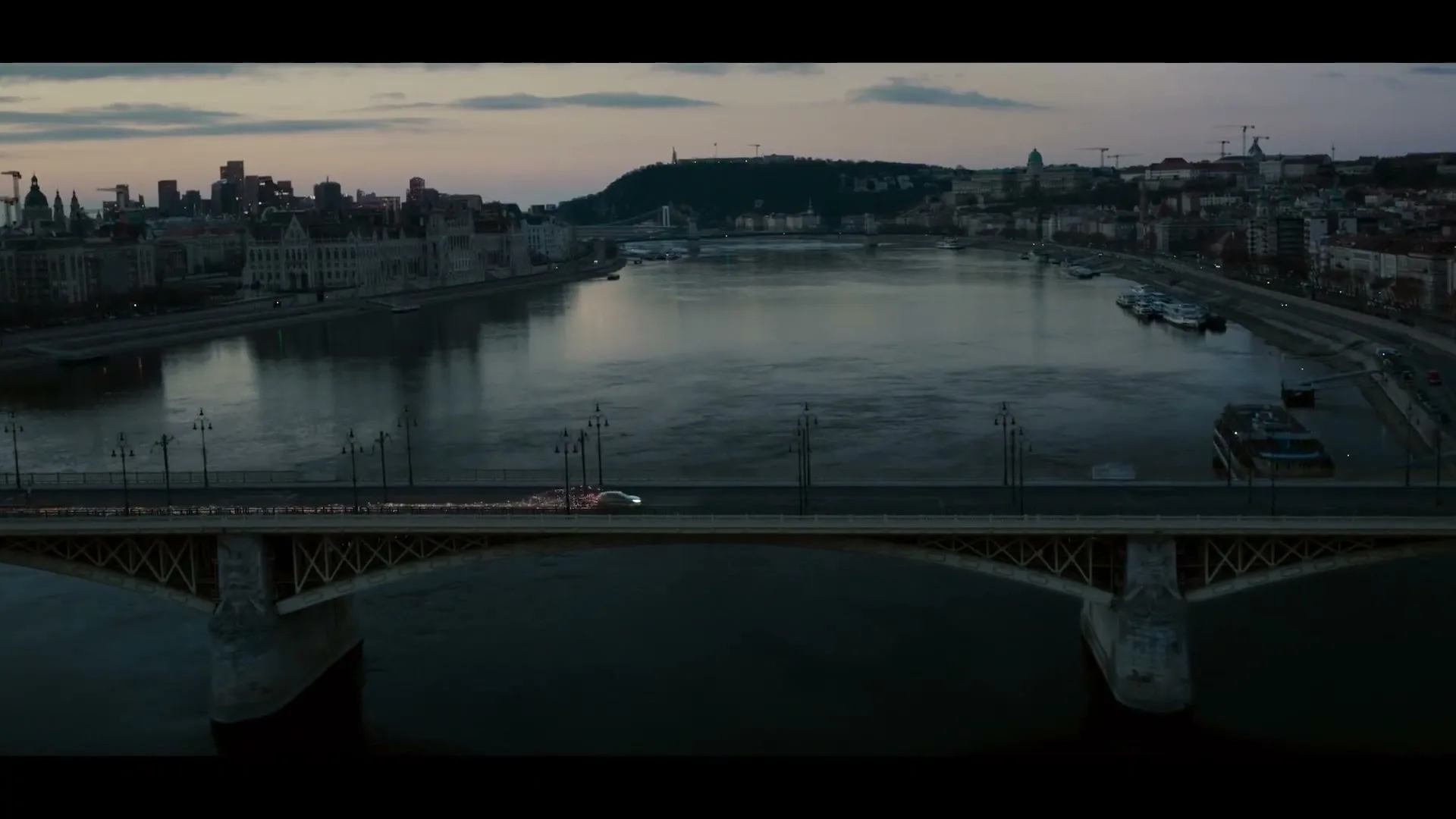 A silver Genesis car drives across a bridge over a river in a European city at dusk, with city lights reflecting on the water.
