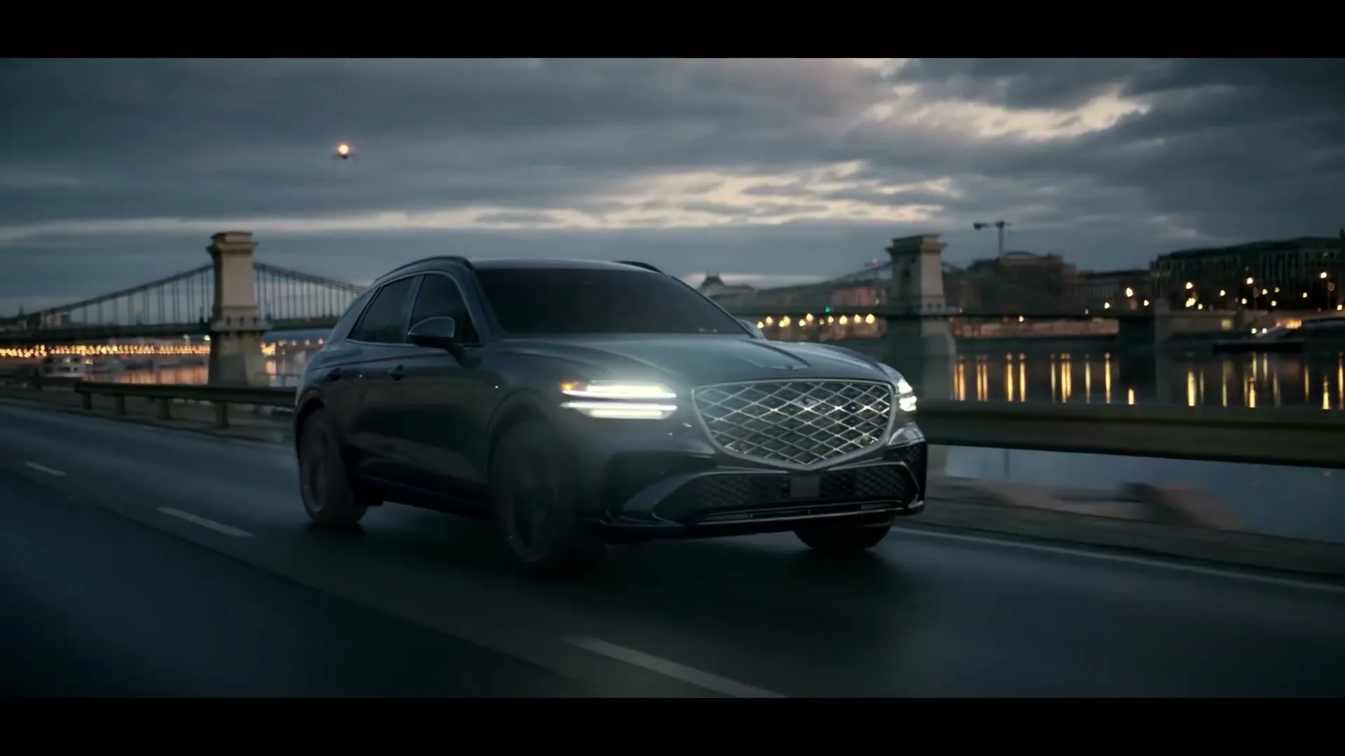 A dark grey Genesis SUV drives on a wet road at dusk, with a lit-up bridge and city lights in the background.