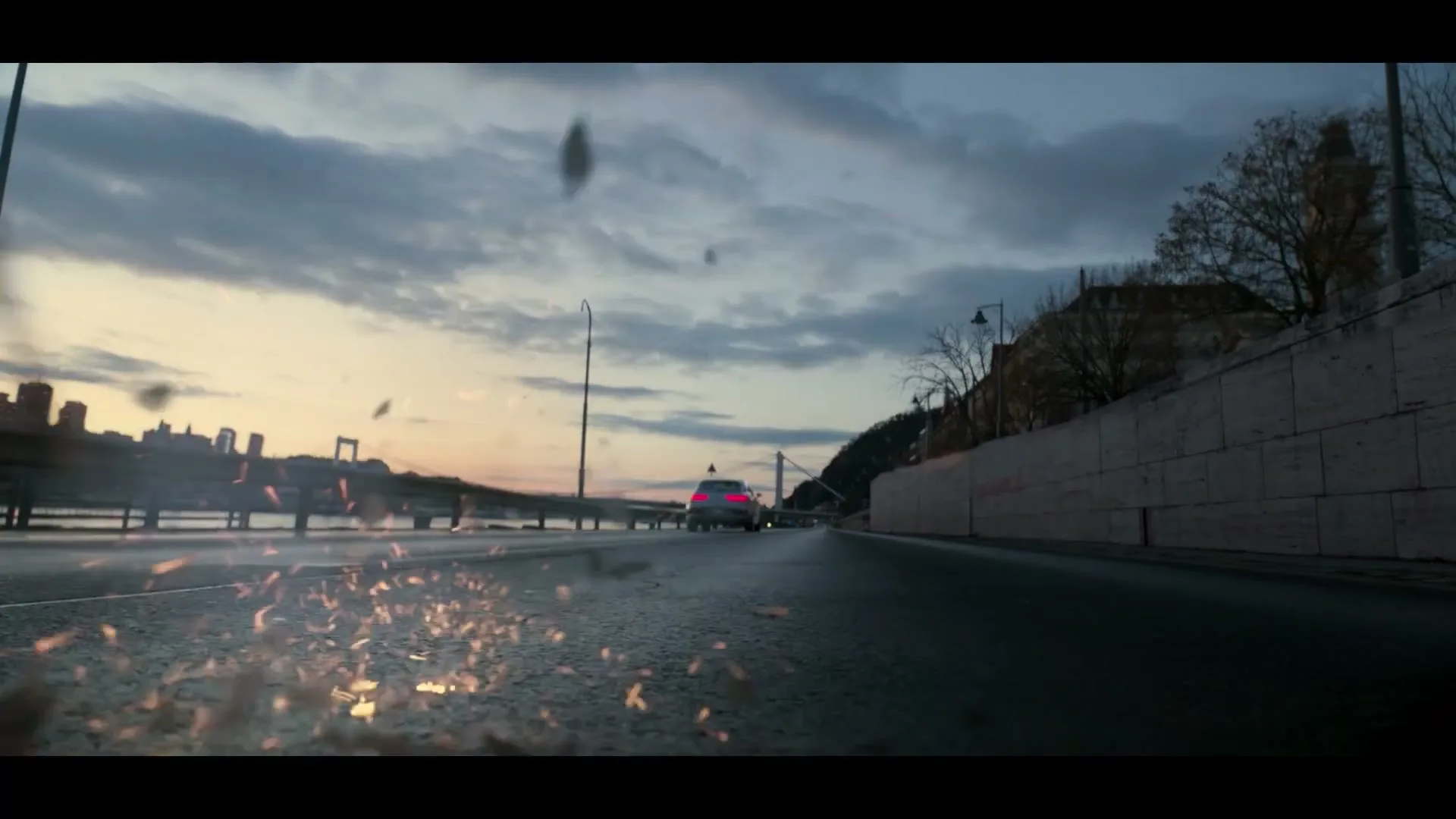 A dark grey Genesis car speeds away on a wet road at dusk, with city buildings and a bridge in the background.