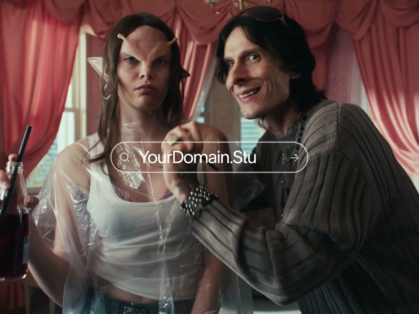 A makeup artist applies prosthetics to a model with elf ears, while another model with blue face paint sits nearby in a pink room.